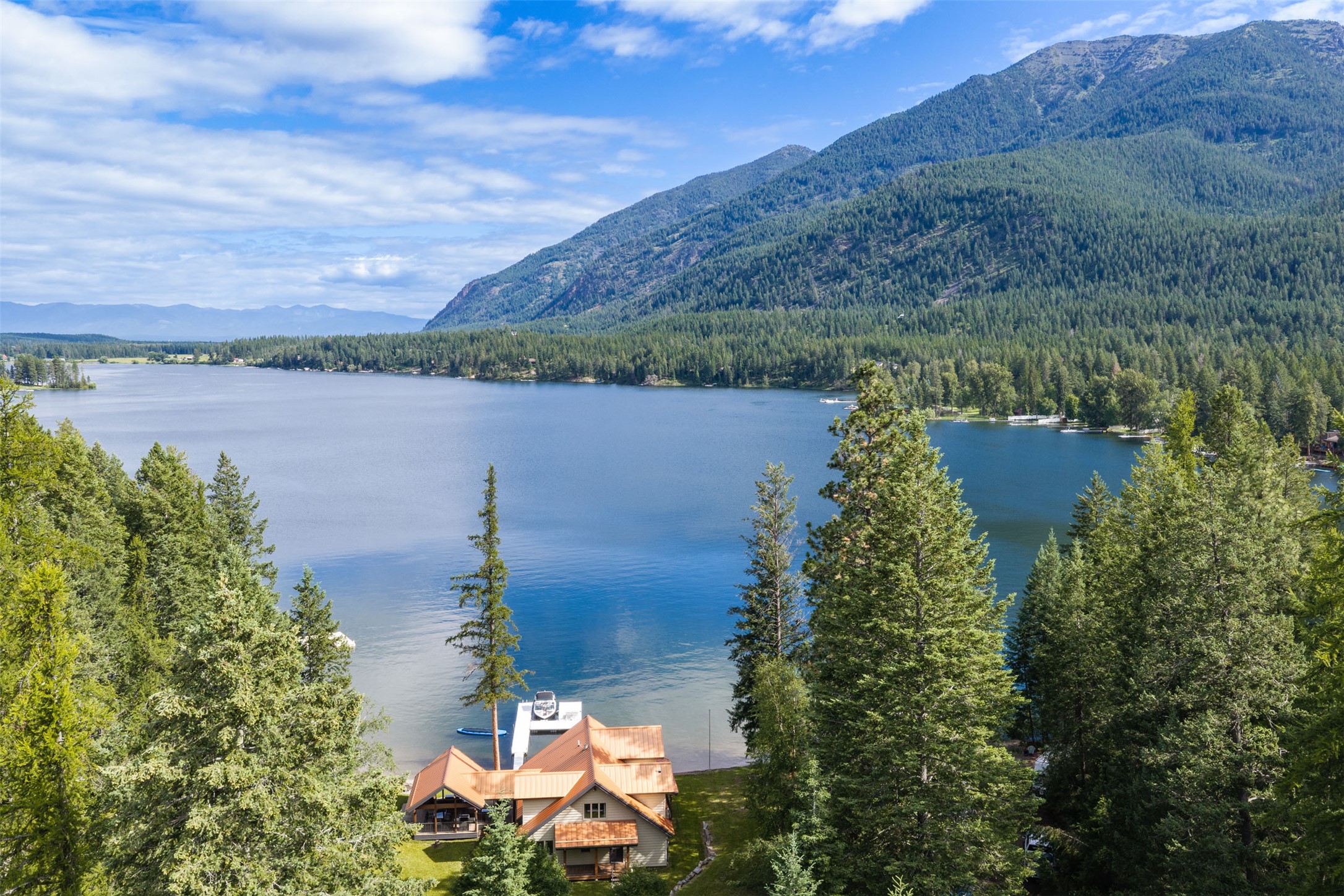 Aerial view of a lakeside home among evergreens, with a lake and forested mountains beyond.
