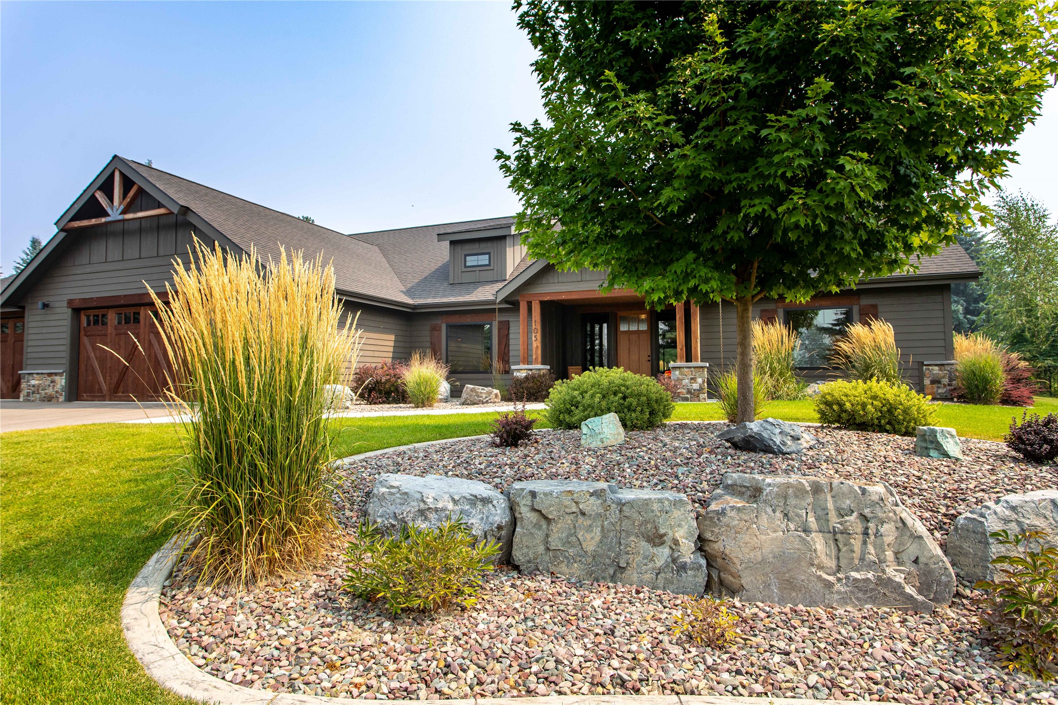 Modern gray home with a wood-trimmed porch, large shade tree, and landscaped rock garden.