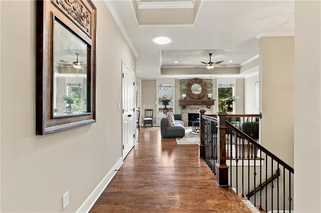 Upper-floor hallway with hardwood floors and dark railing overlooking a furnished living room with a fireplace.