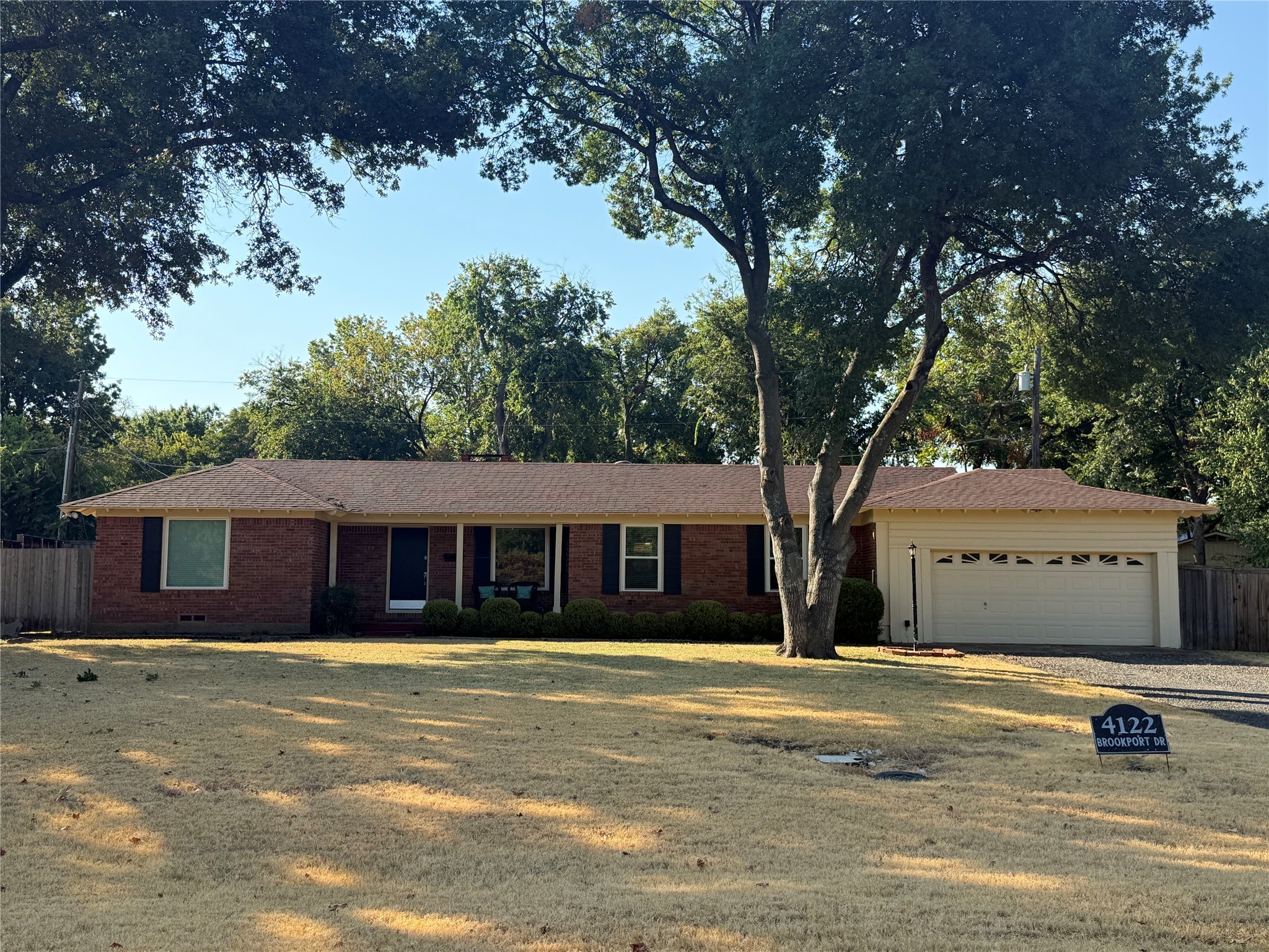 Single-story brick house with an attached garage, shaded by a large tree; yard sign reads 4122 Brookport Dr.