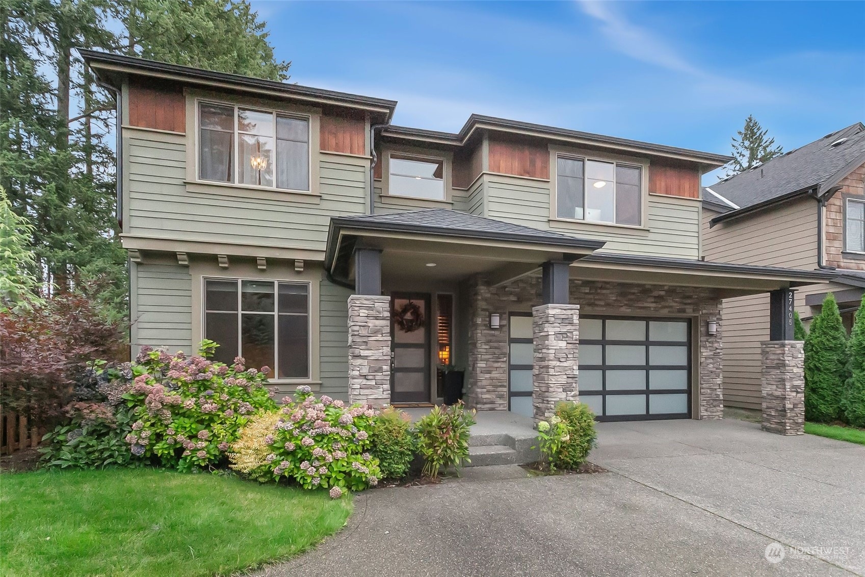Two-story sage-green house with stone porch columns, a frosted-panel garage door, and flowering front-yard shrubs.