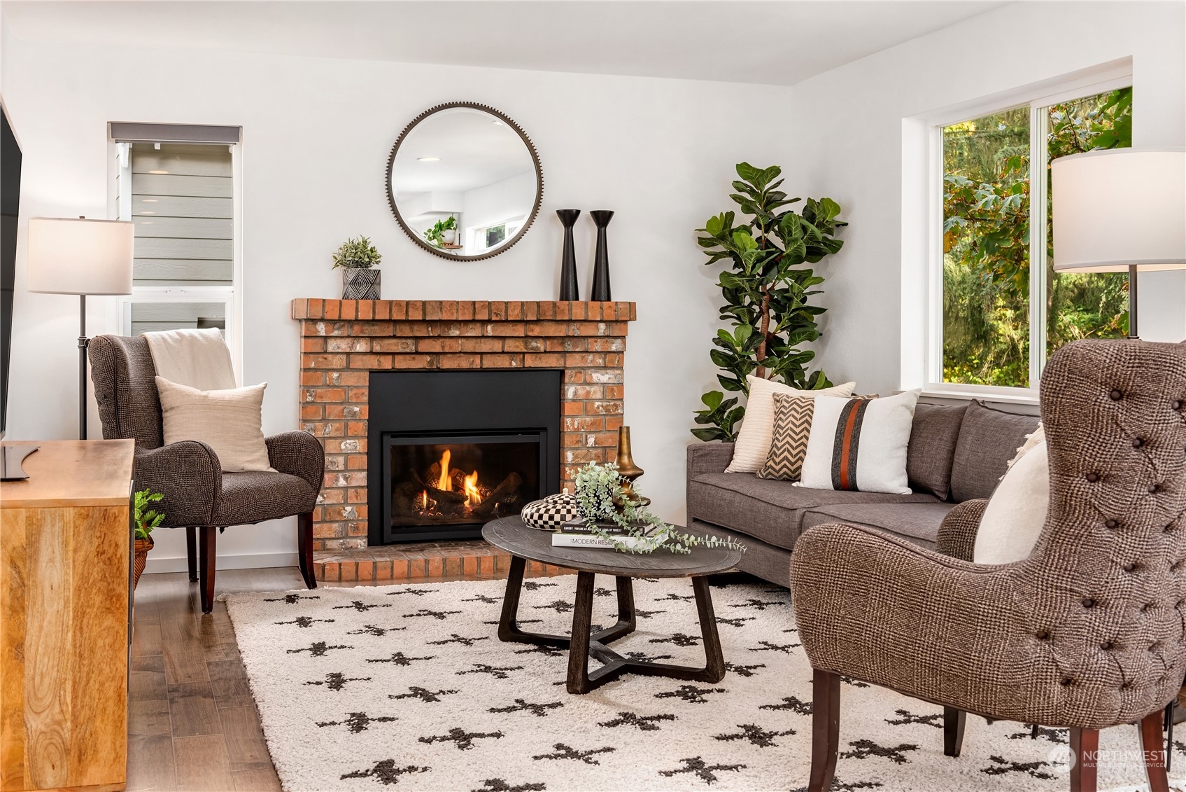 Staged living room with a brick fireplace, lit fire, gray sofa, and large window overlooking trees.
