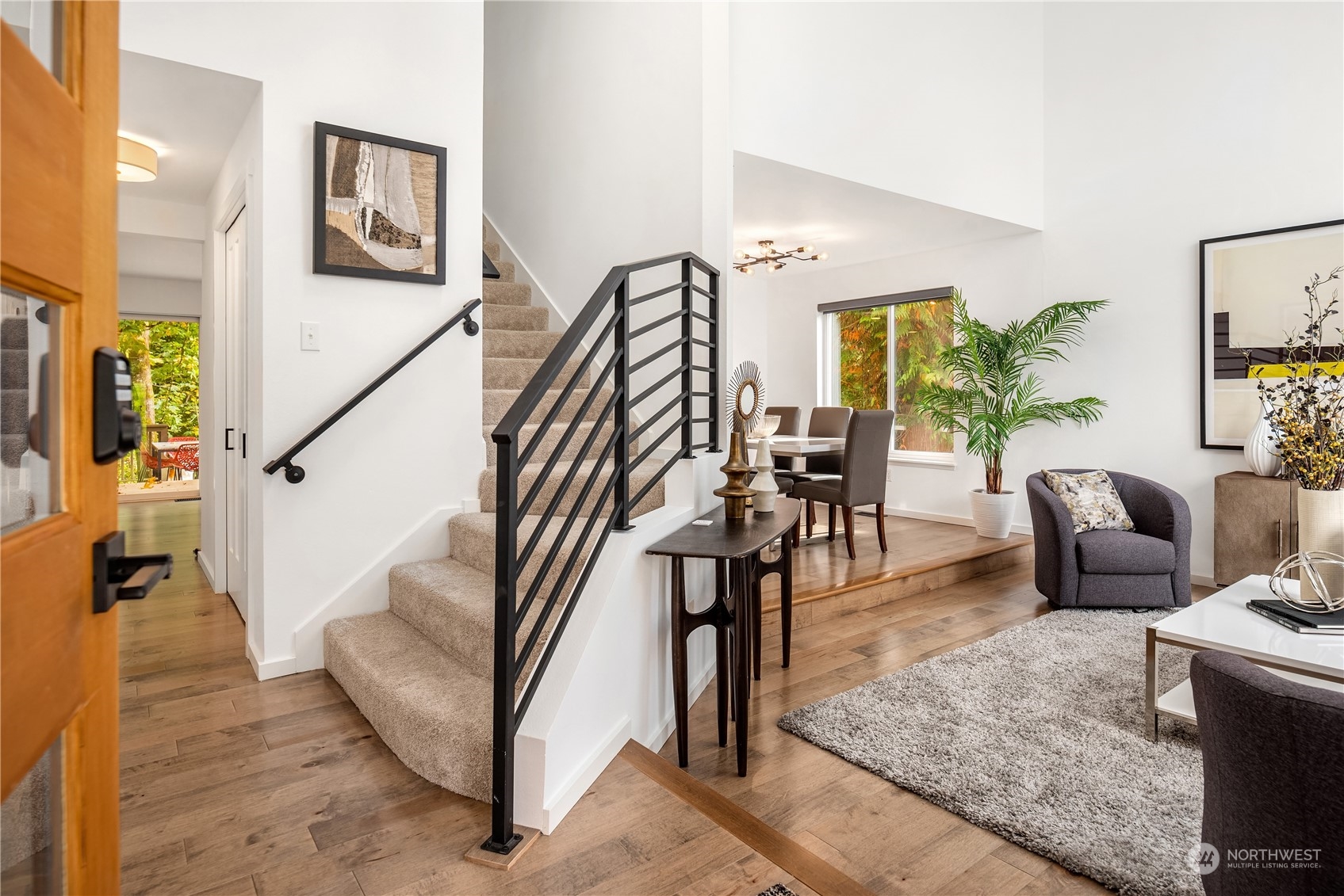 Bright entry and living room with carpeted stairs, black railings, wood floors, and a dining area.