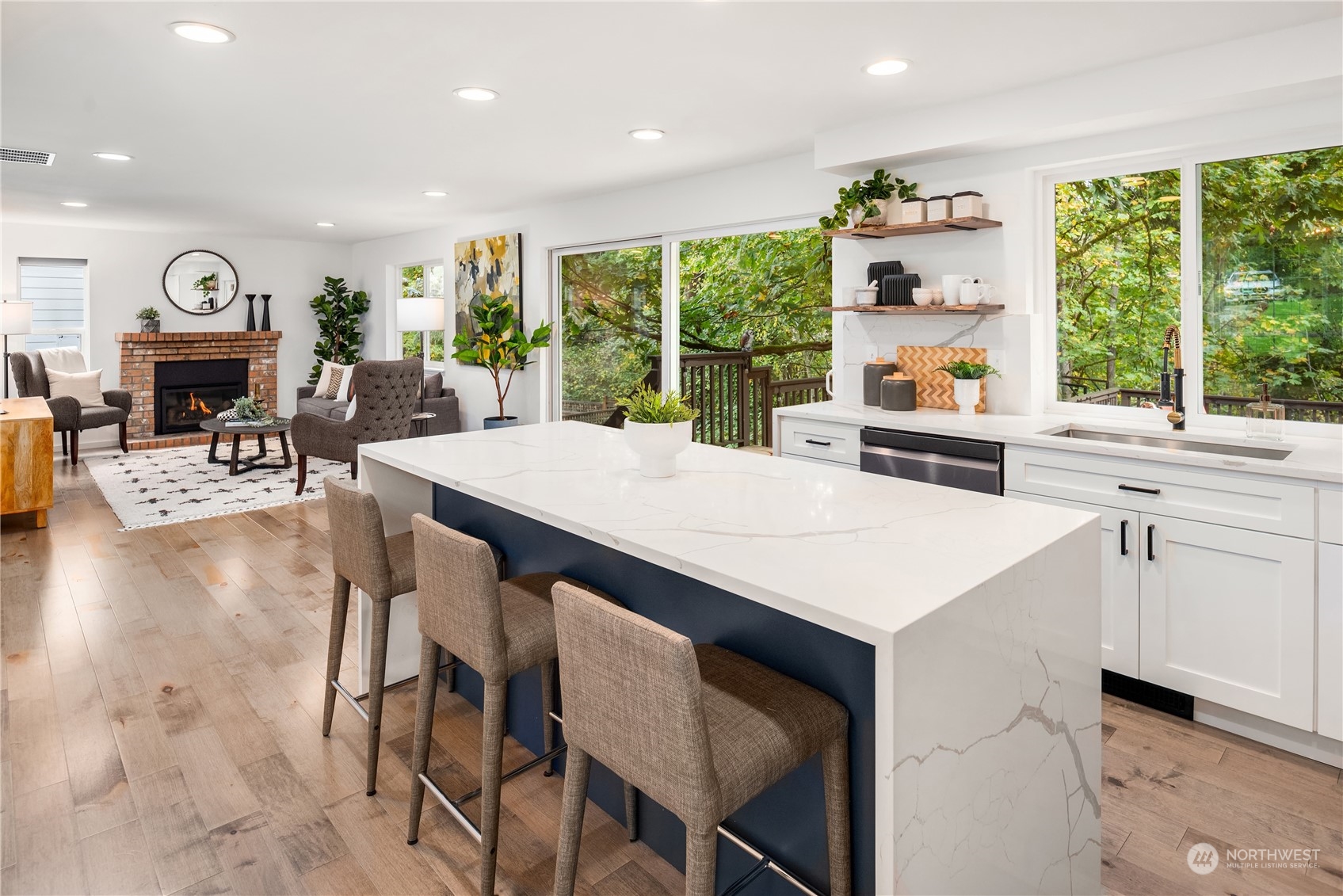 Open kitchen and living room with a marble-look island, navy panel, and leafy views through large windows.
