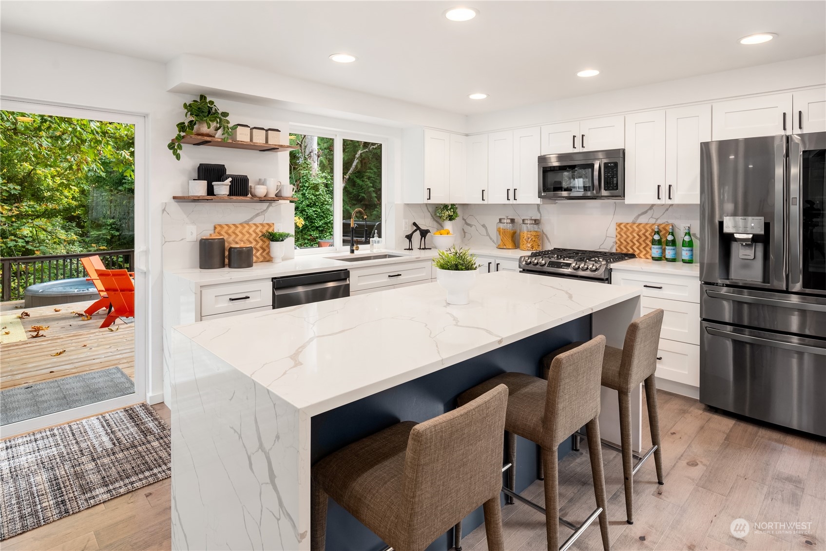 Bright kitchen with a large waterfall island, white cabinets, stainless appliances, and a deck visible through glass doors.