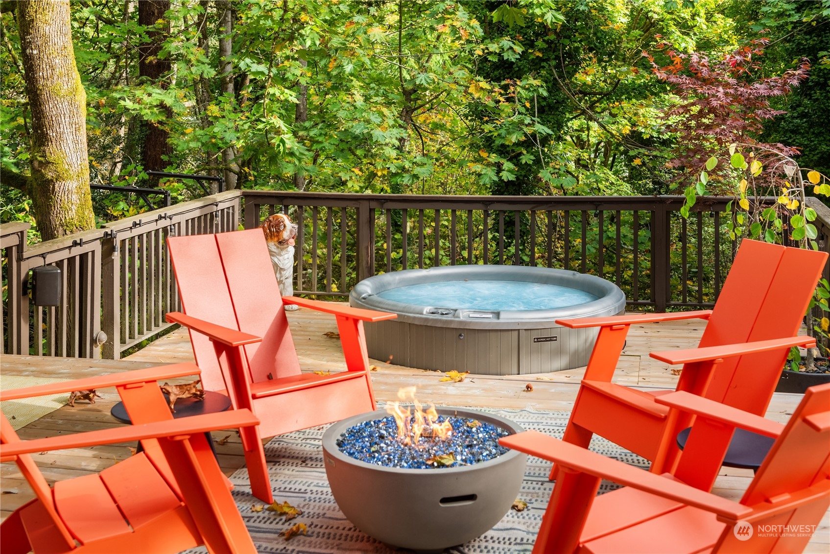 Wooden deck with a hot tub, orange Adirondack chairs, and a fire pit among leafy trees.