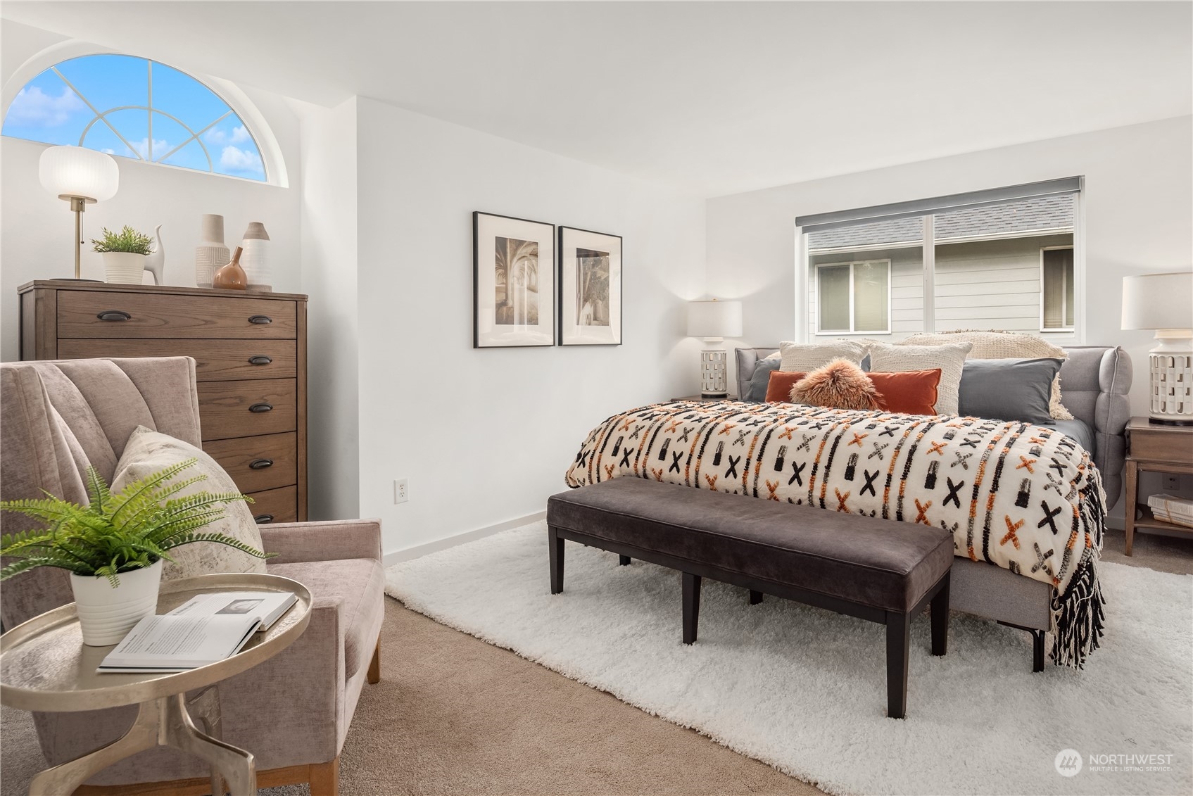 Staged bedroom with a gray bed, patterned blanket, dark bench, and large arched window.