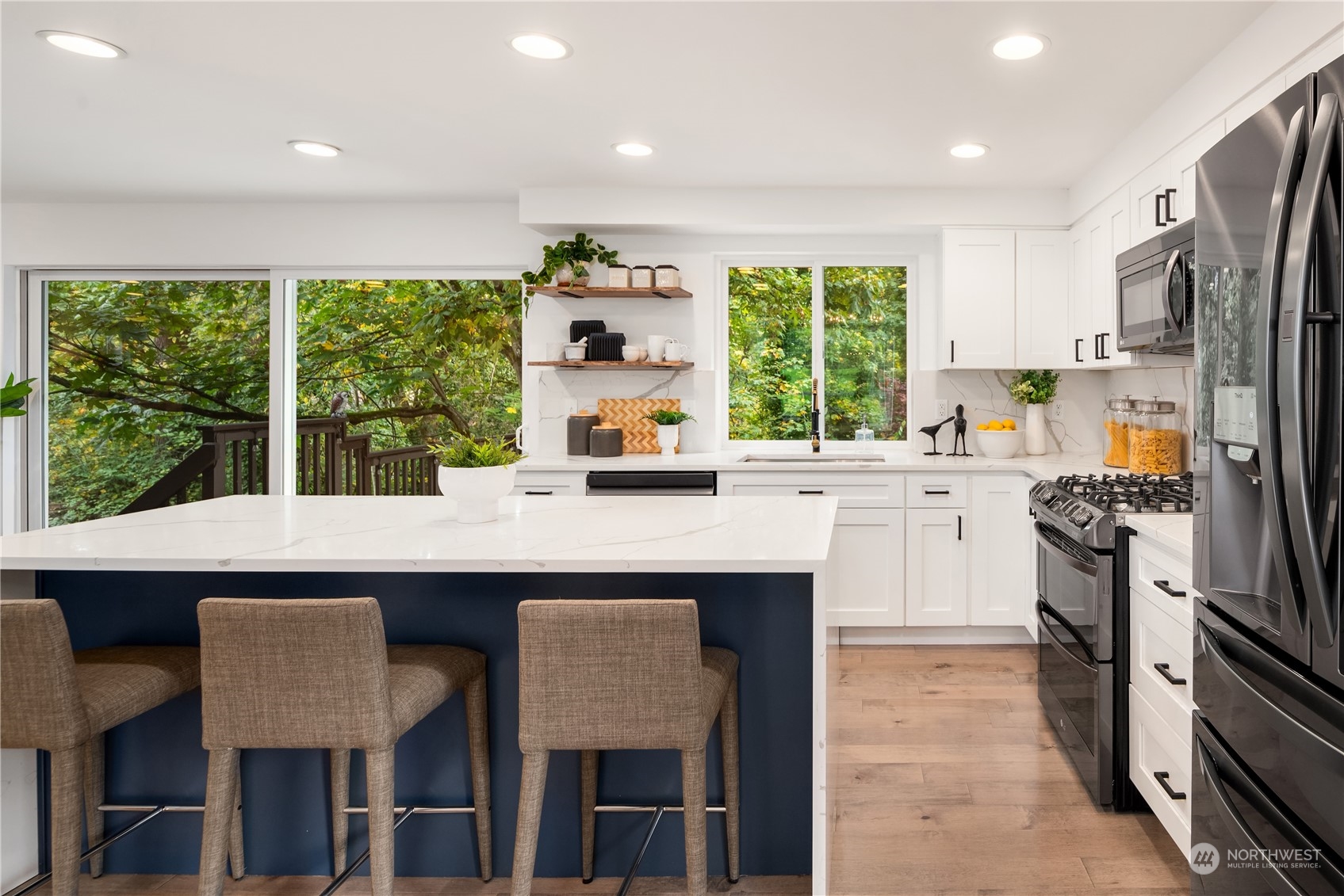 Bright white kitchen with a navy island, bar stools, stainless appliances, and windows overlooking trees.