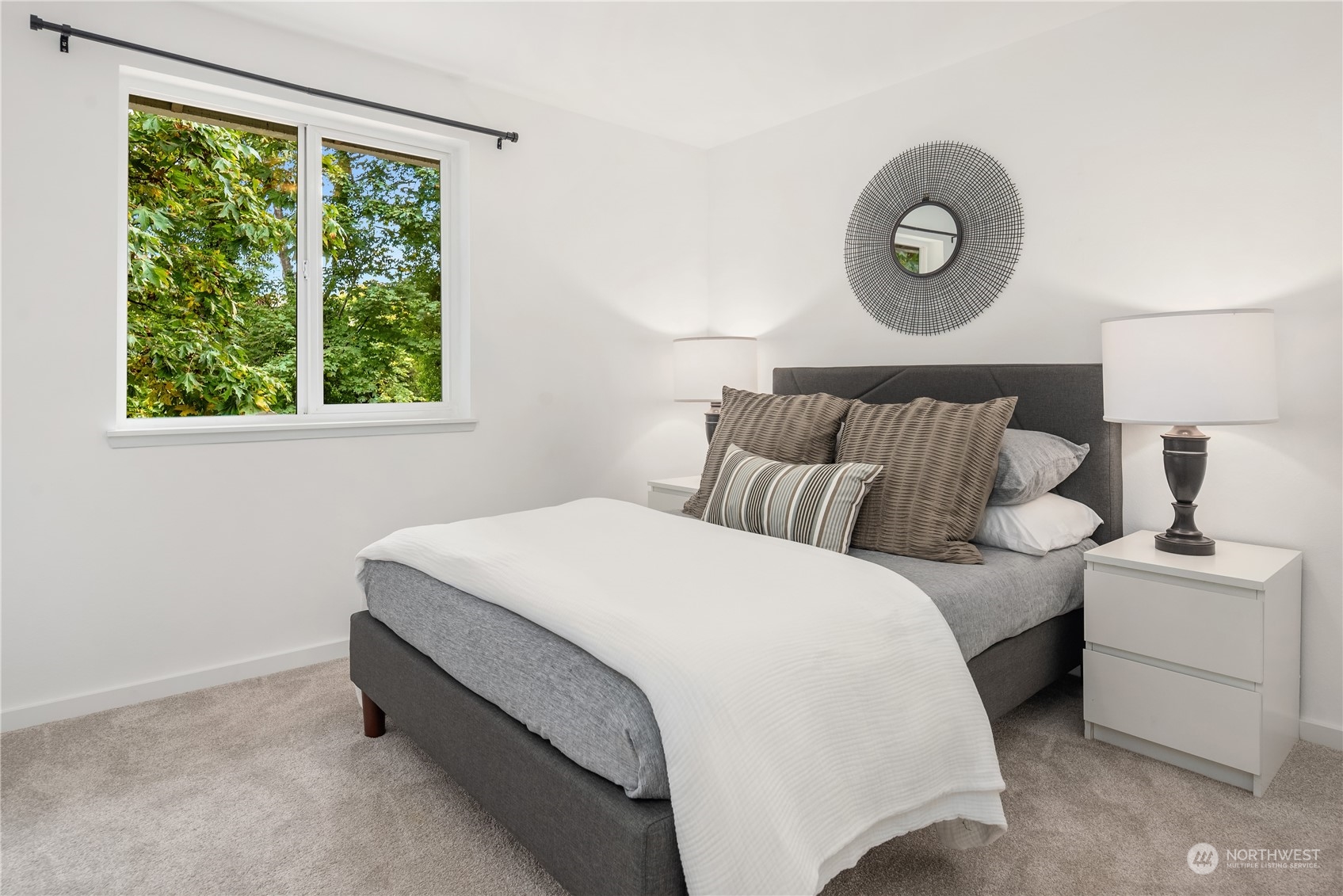 Staged bedroom with a gray bed, white bedding, and a large window overlooking leafy trees.