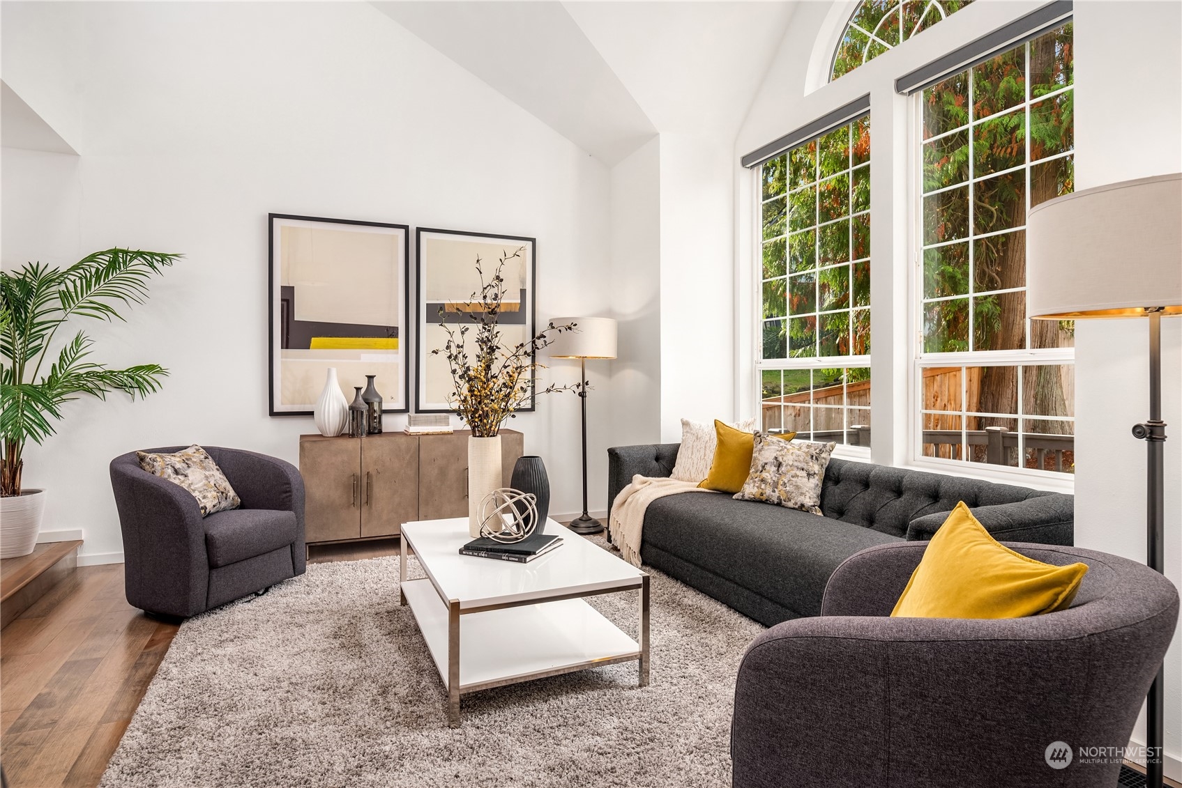 Staged living room with vaulted ceiling, gray seating, and tall windows overlooking trees.