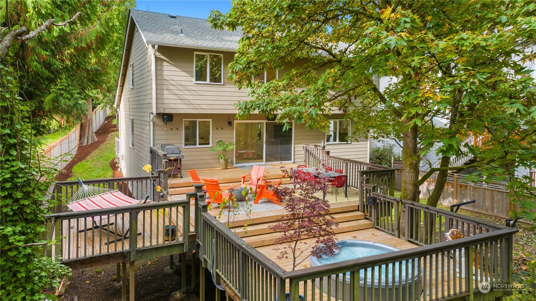 Rear of a beige house with a tiered deck, outdoor seating, hammock, and hot tub beneath mature trees.