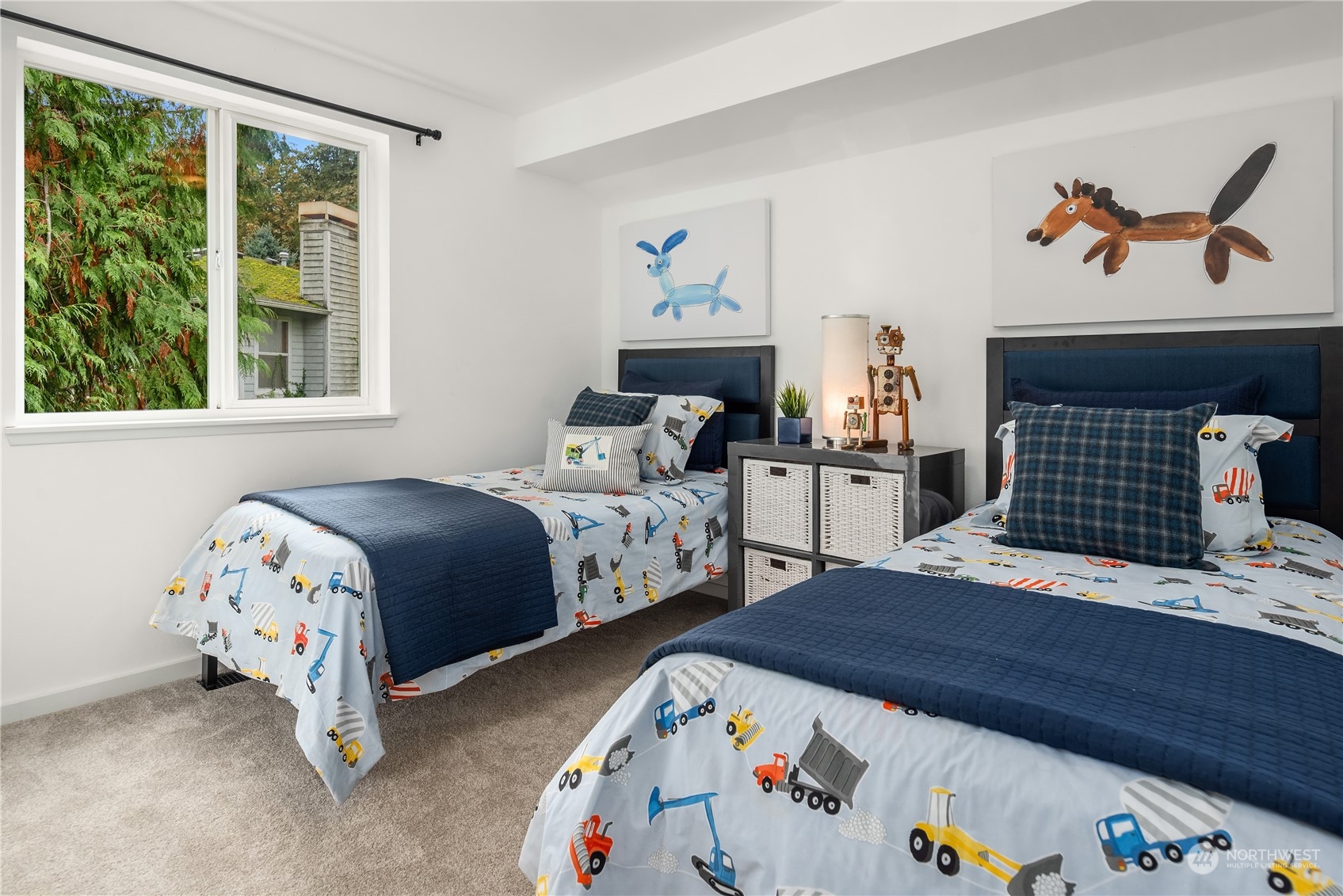 Bedroom with two twin beds, construction-vehicle bedding, and a large window overlooking leafy trees.