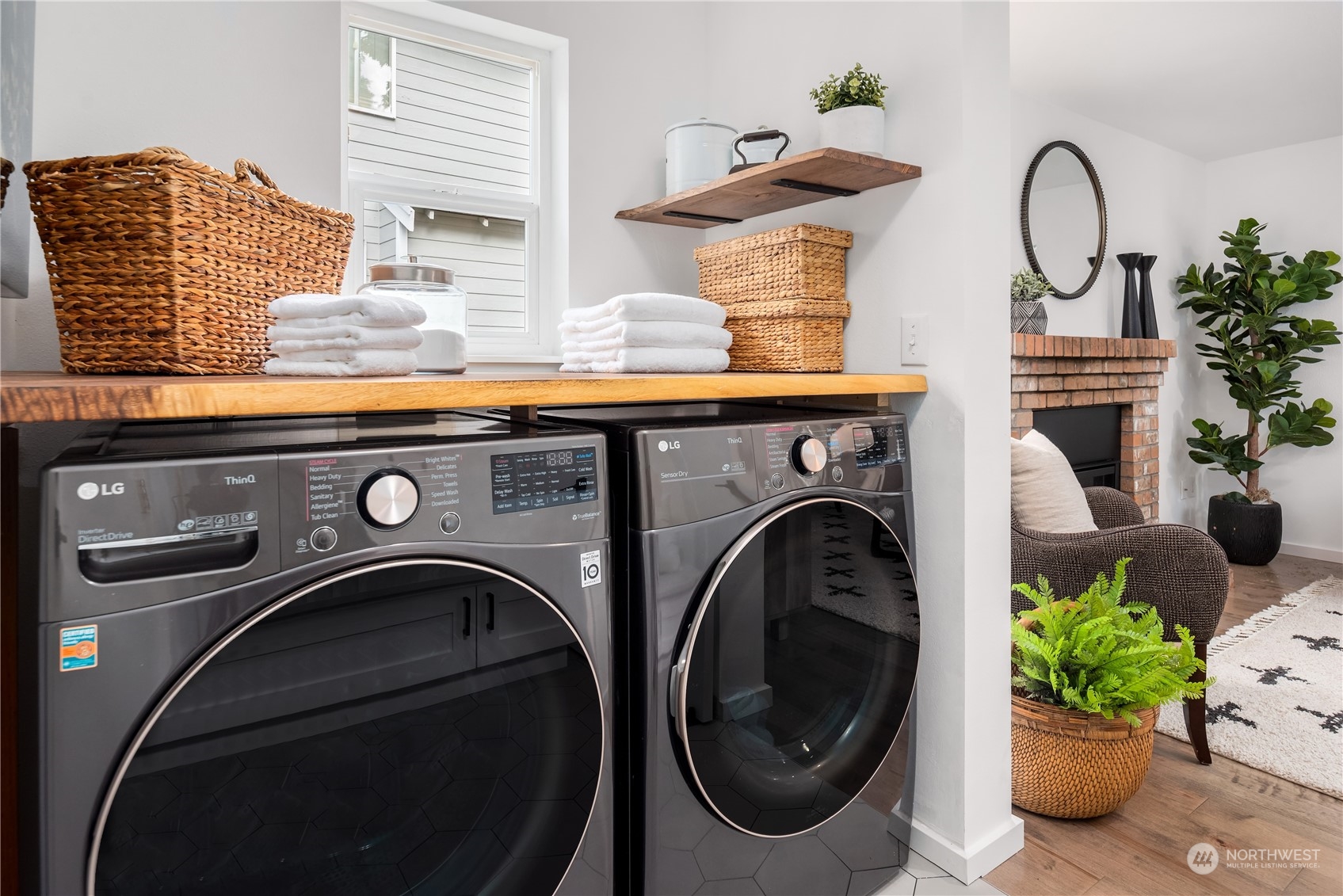 Laundry room with front-loading washer and dryer, wood counter, woven baskets, and an adjoining brick fireplace.