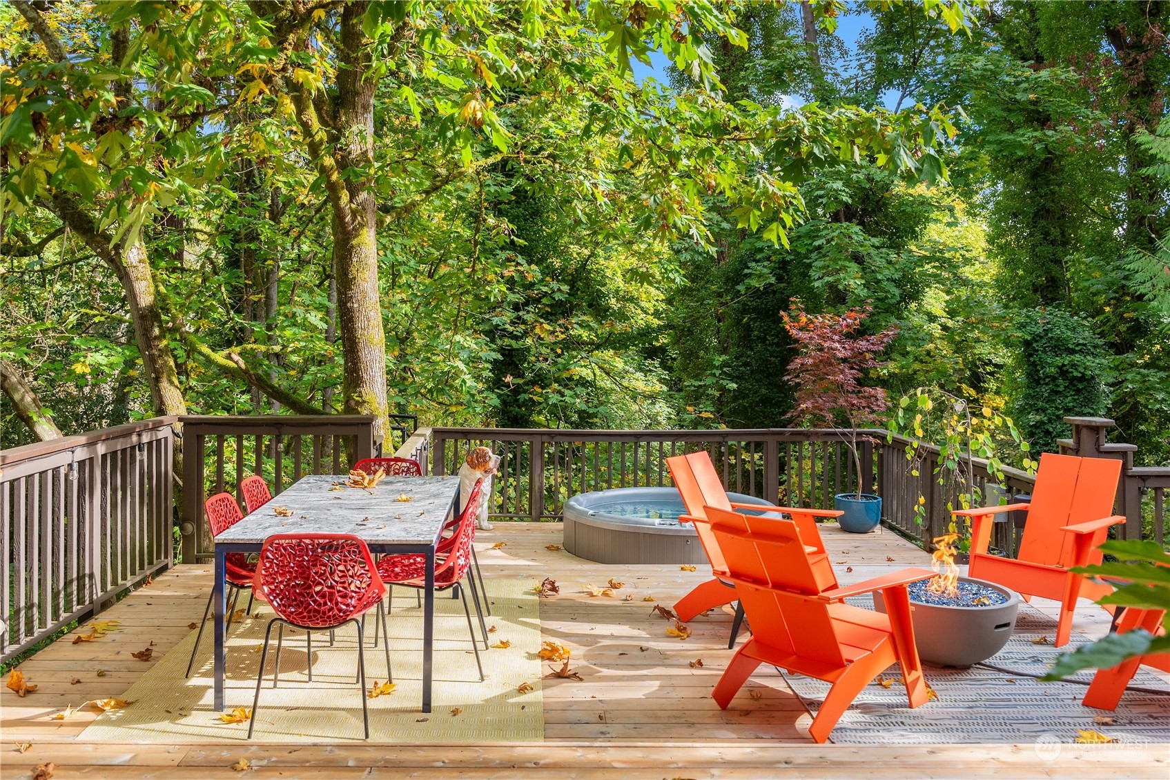 Wooden deck with a hot tub, fire pit, orange lounge chairs, and wooded surroundings.