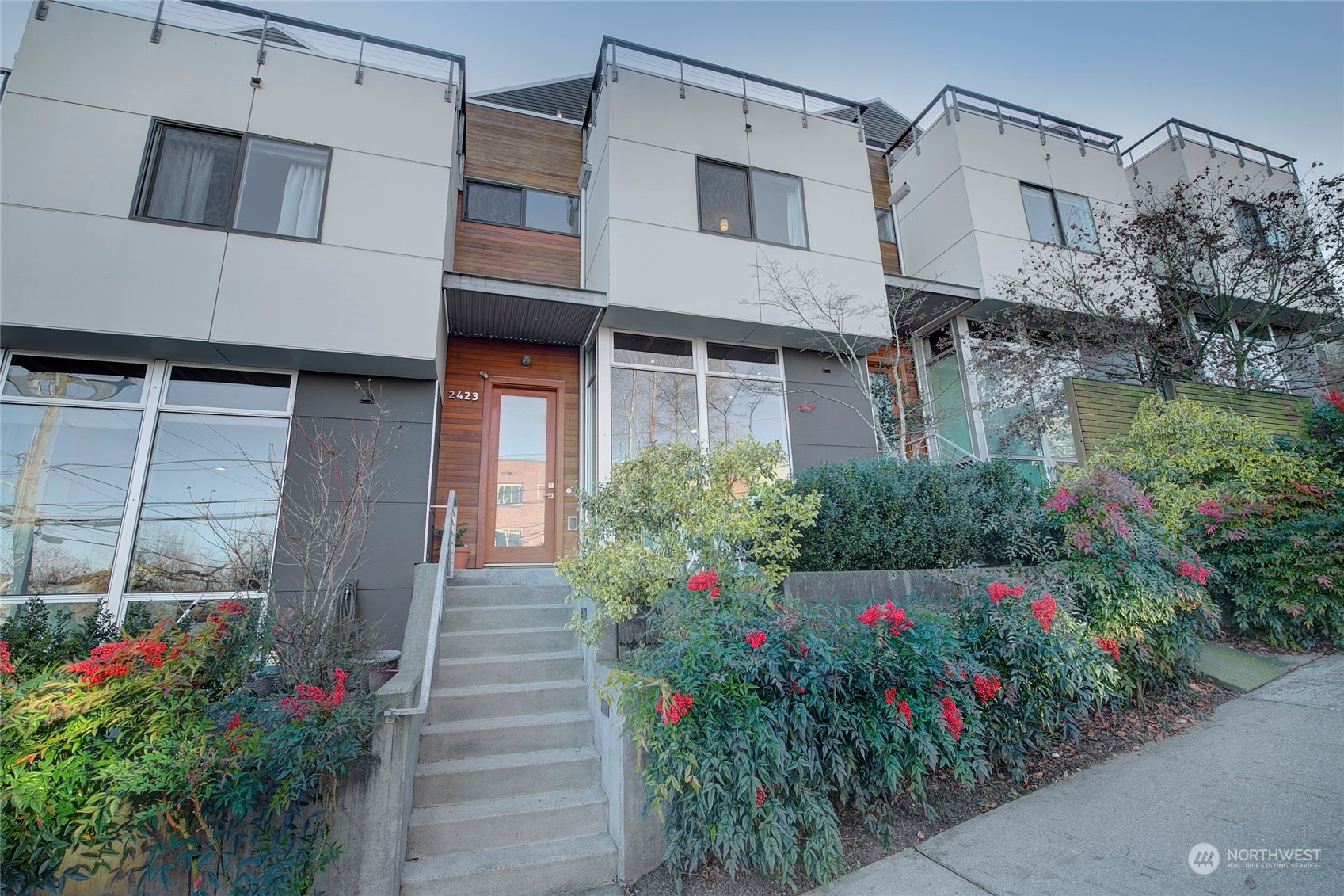Modern townhouse with wood-trimmed entry, rooftop railings, concrete steps, and flowering shrubs.