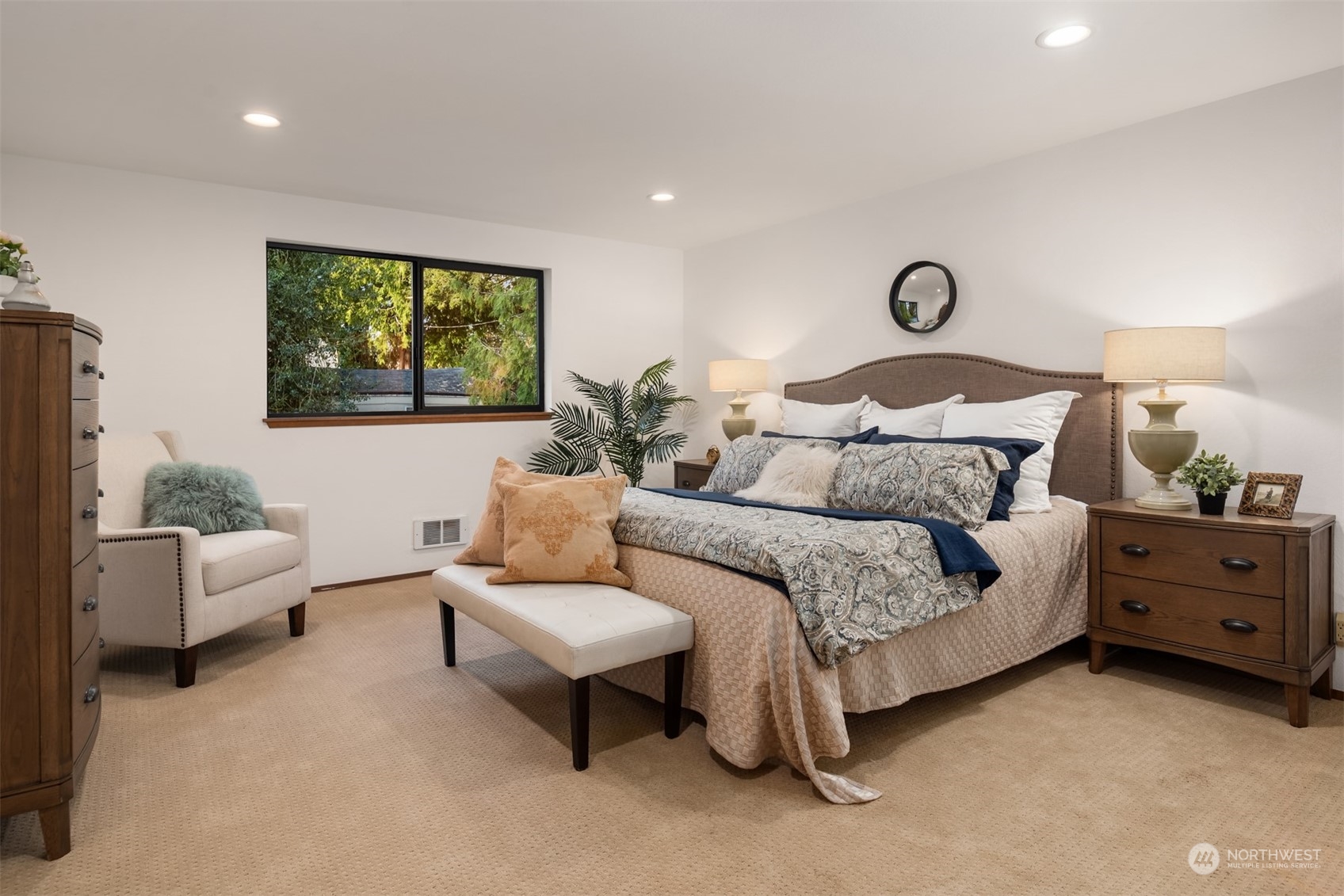 Furnished bedroom with a large patterned bed, cushioned bench, armchair, and wide window overlooking trees.