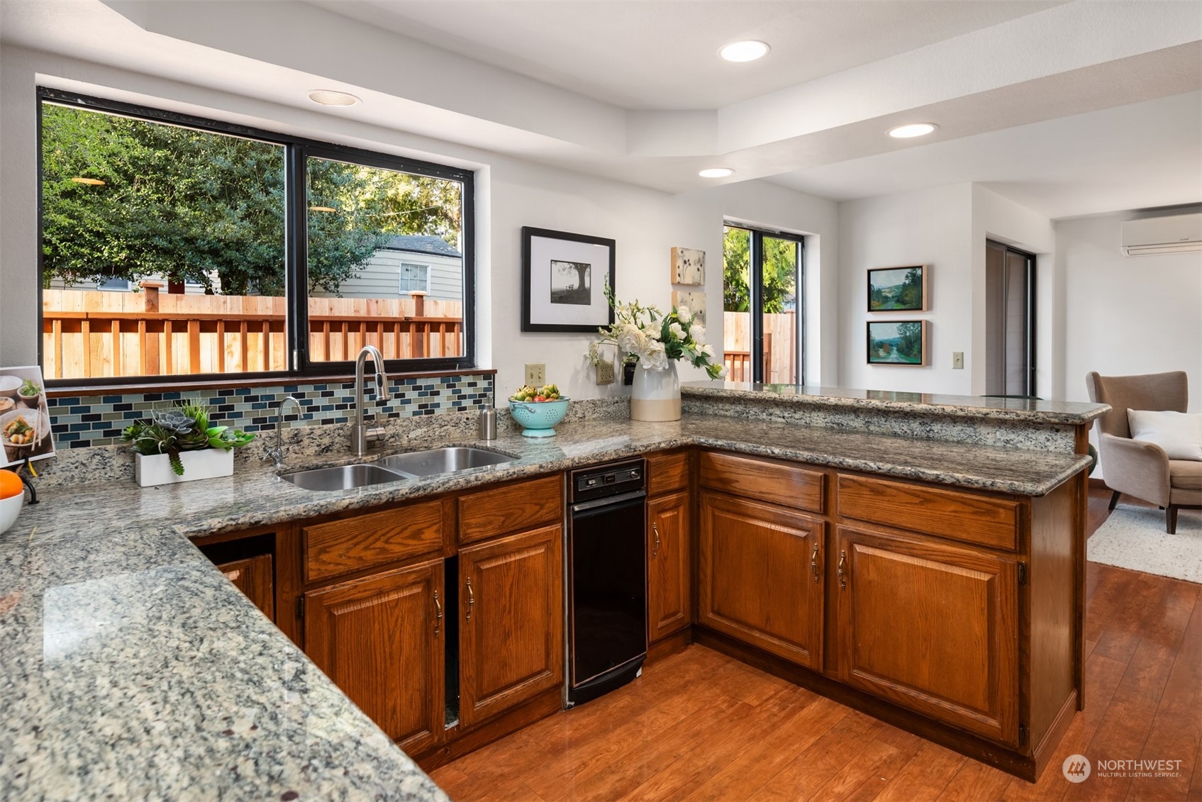 Kitchen with granite counters, wood cabinets, colorful tile backsplash, and large windows overlooking a fenced yard.