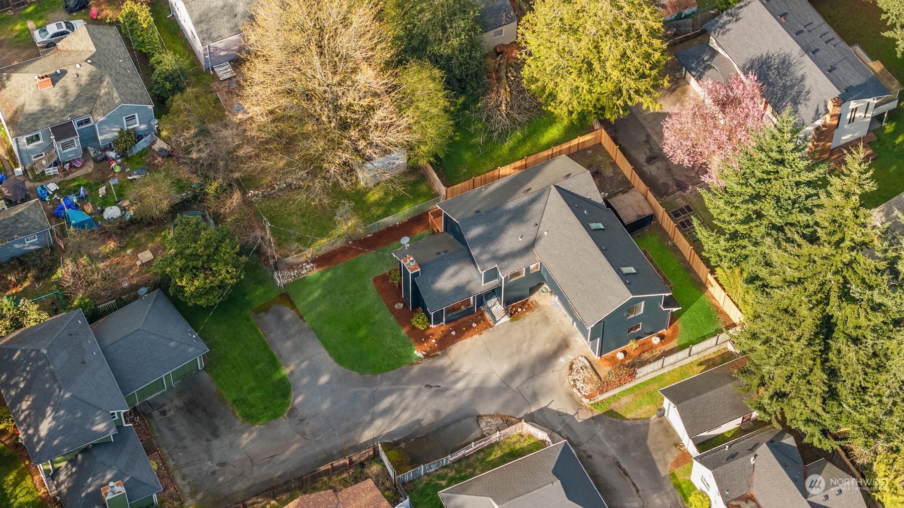Aerial view of a dark-sided home with a fenced yard, broad driveway, and autumn trees.