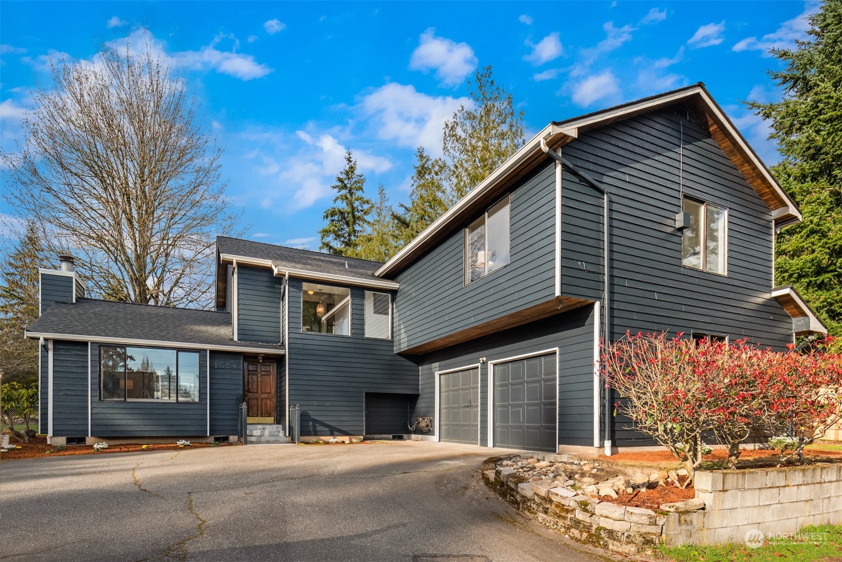 Dark-sided two-story house with an attached garage, broad driveway, and landscaped stone retaining wall.