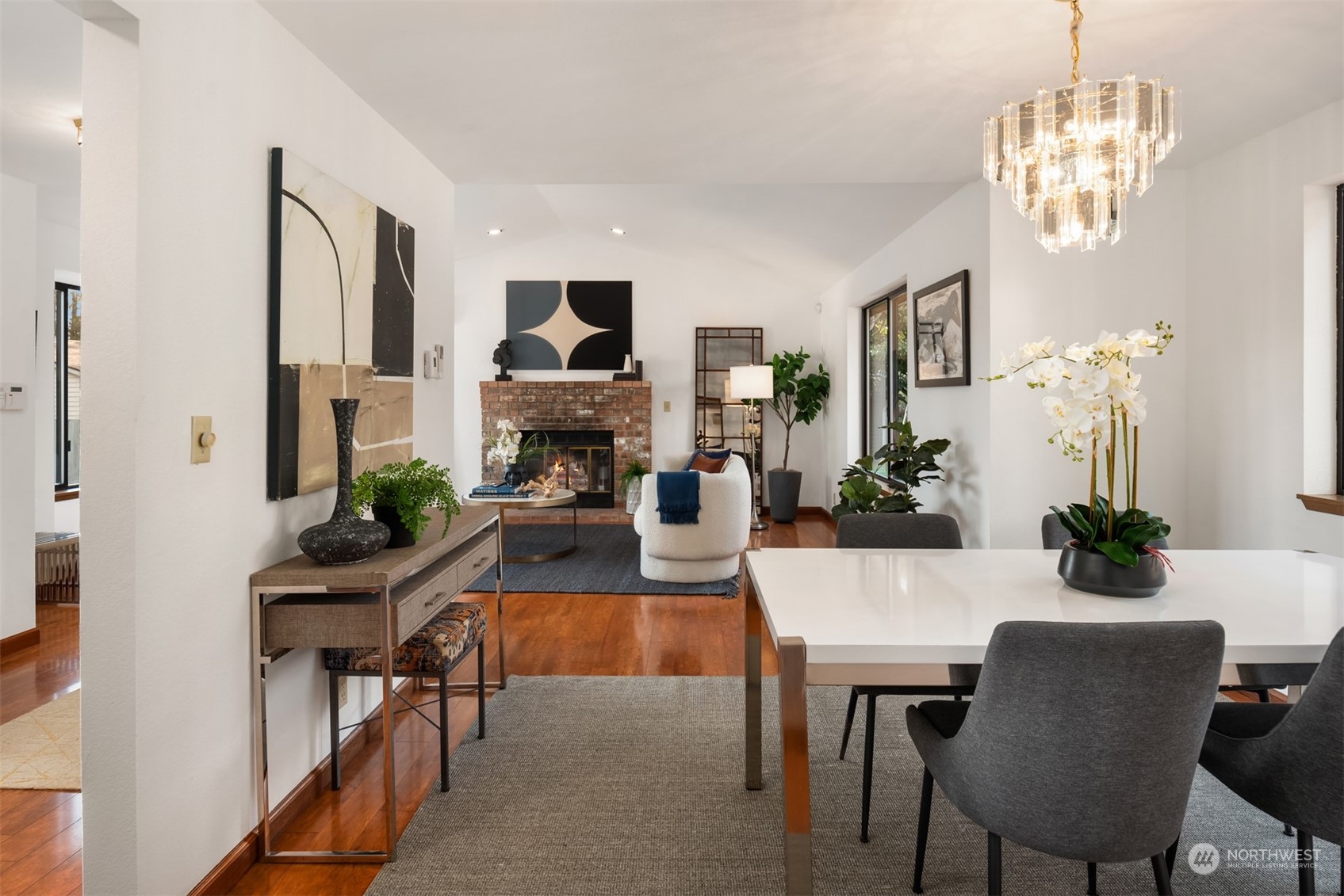 Open-plan living and dining room with a brick fireplace, polished wood floors, and a glass chandelier.