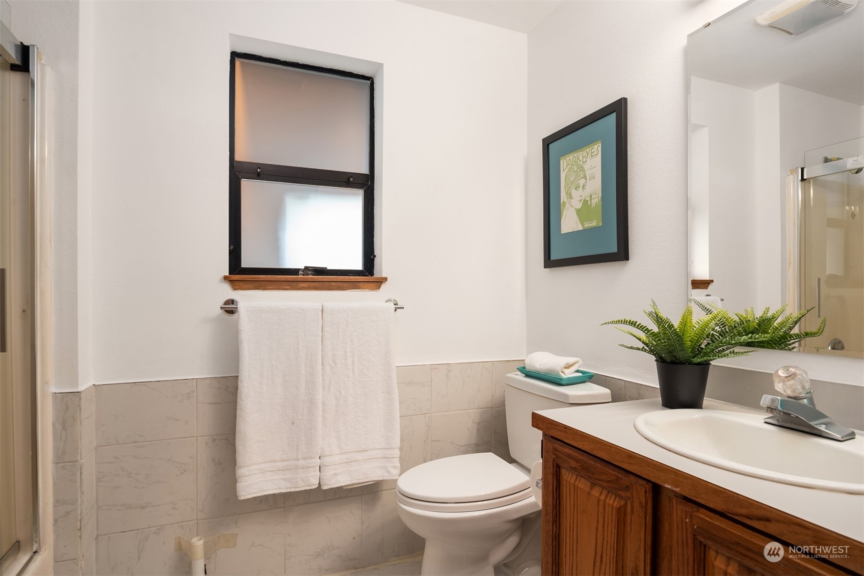 Bathroom with marble-look tile, a wood vanity, black-framed window, and potted fern.