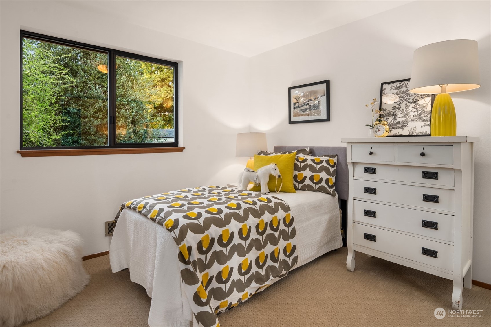 Bedroom with a patterned bed, white dresser, and large window overlooking greenery.