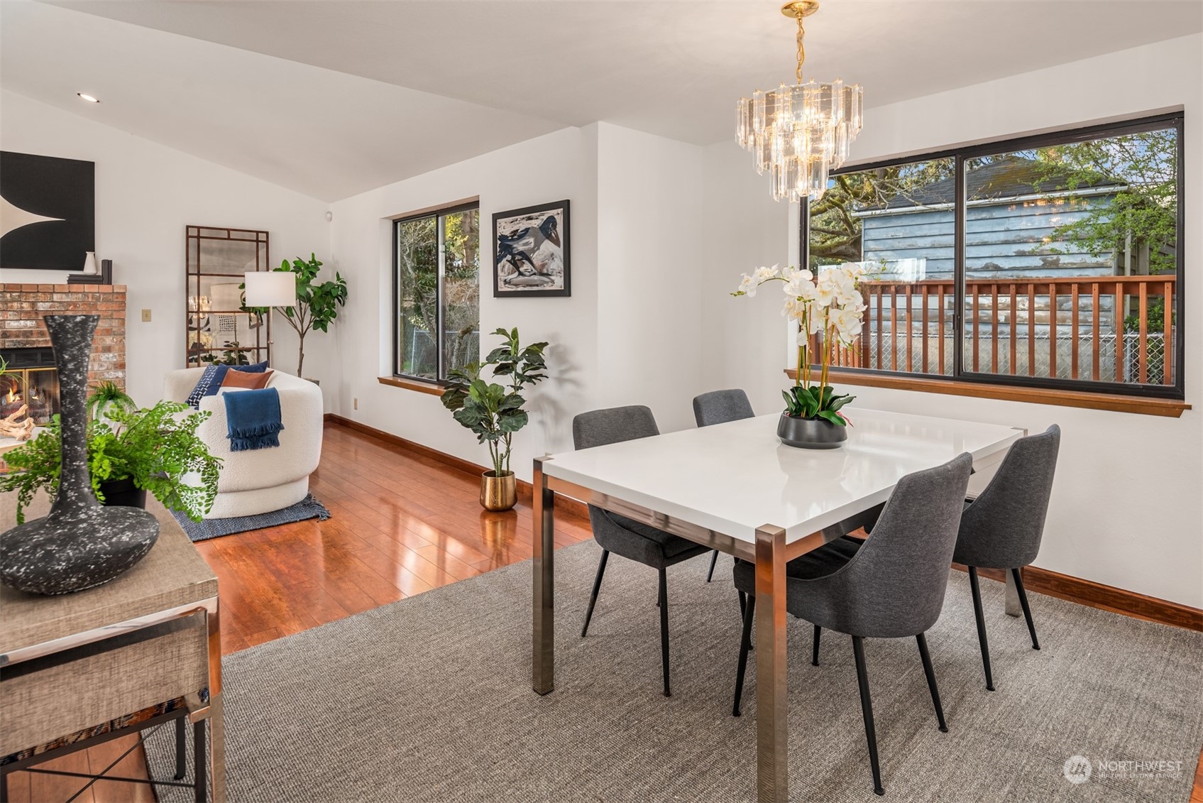 Dining and living area with a brick fireplace, white dining table, hardwood floors, and large deck-facing windows.