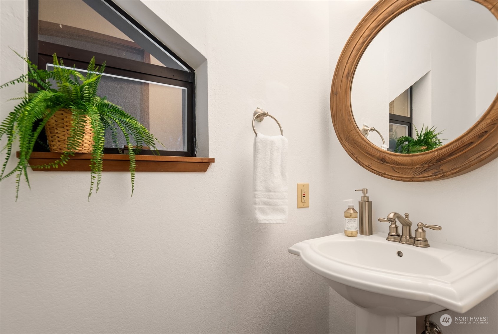 Bathroom vanity with a round wood-framed mirror and fern beside the window.