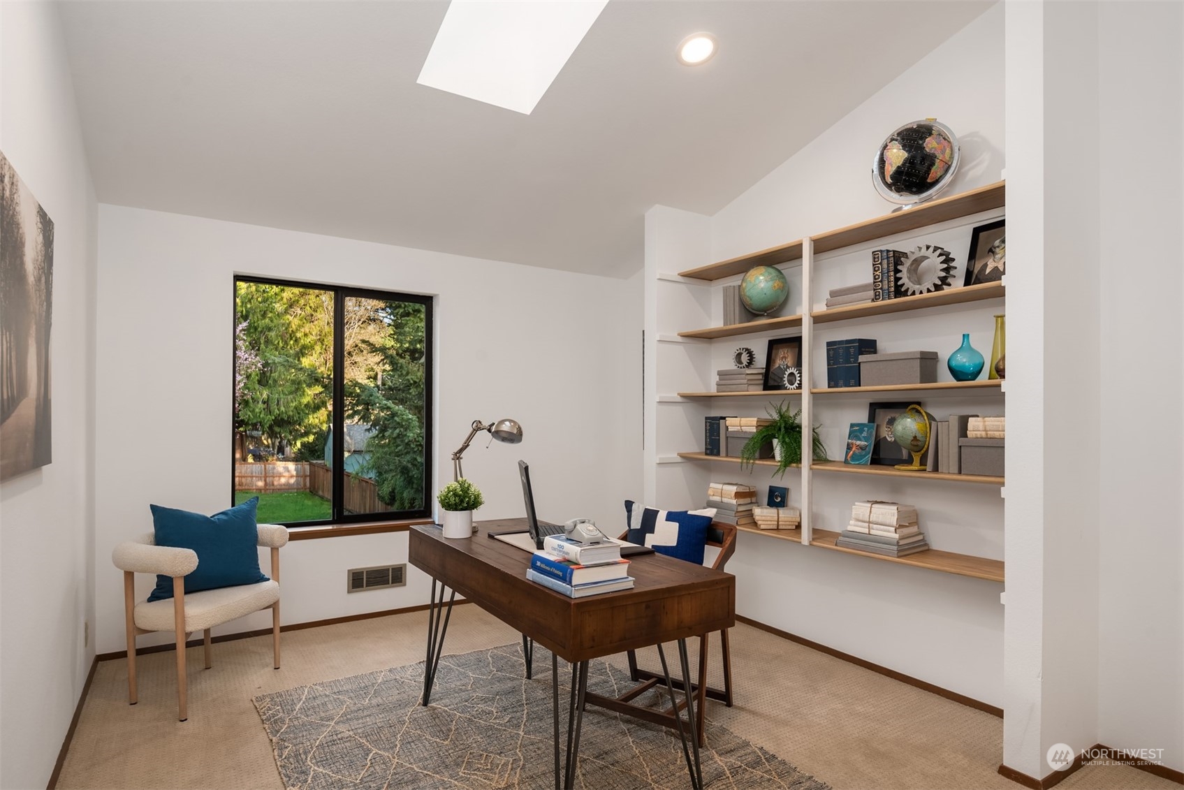Furnished home office with built-in shelves, a dark wood desk, and a large window overlooking the yard.