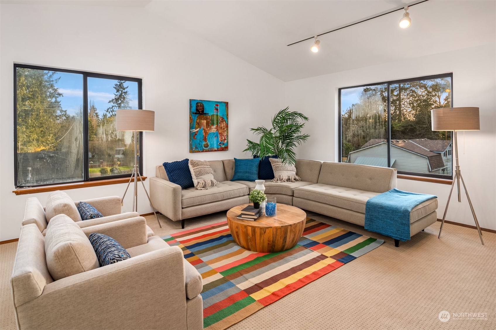 Staged living room with a beige sectional, colorful rug, and large windows overlooking trees and rooftops.