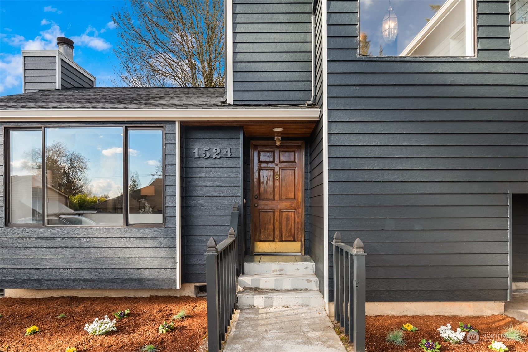 Home entrance with dark gray siding, a wooden door, concrete steps, and landscaped flower beds.