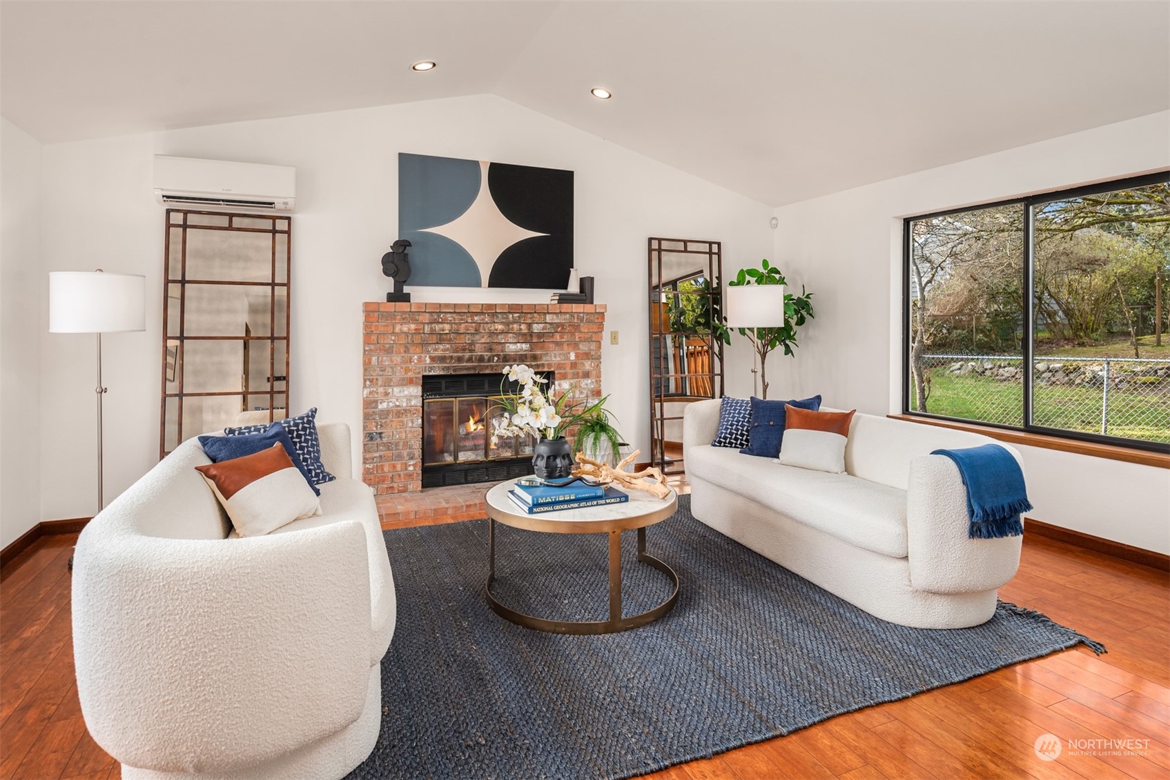 Staged living room with a brick fireplace, white sofas, and large windows overlooking a fenced yard.