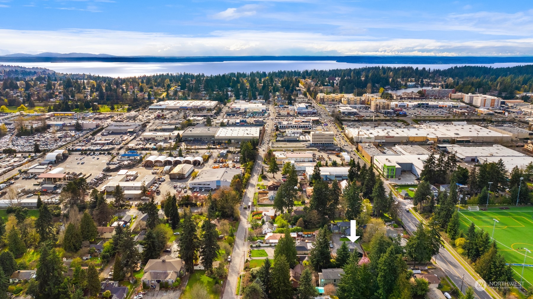 Aerial view of the neighborhood, with a white arrow marking the home amid trees and nearby commercial buildings.