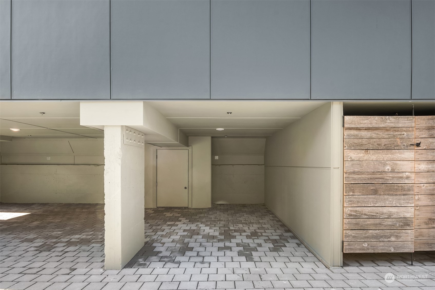 Covered parking bay with gray pavers, a concrete support column, and wood siding.