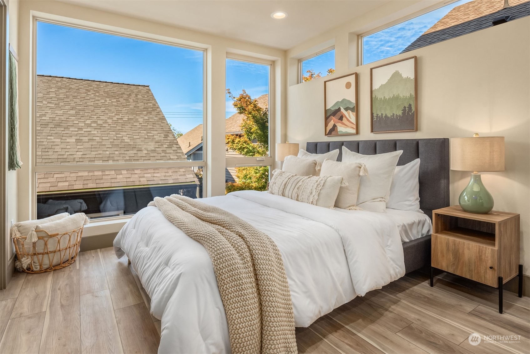 Staged bedroom with a white bed, knit throw, and tall windows overlooking rooftops and autumn trees.