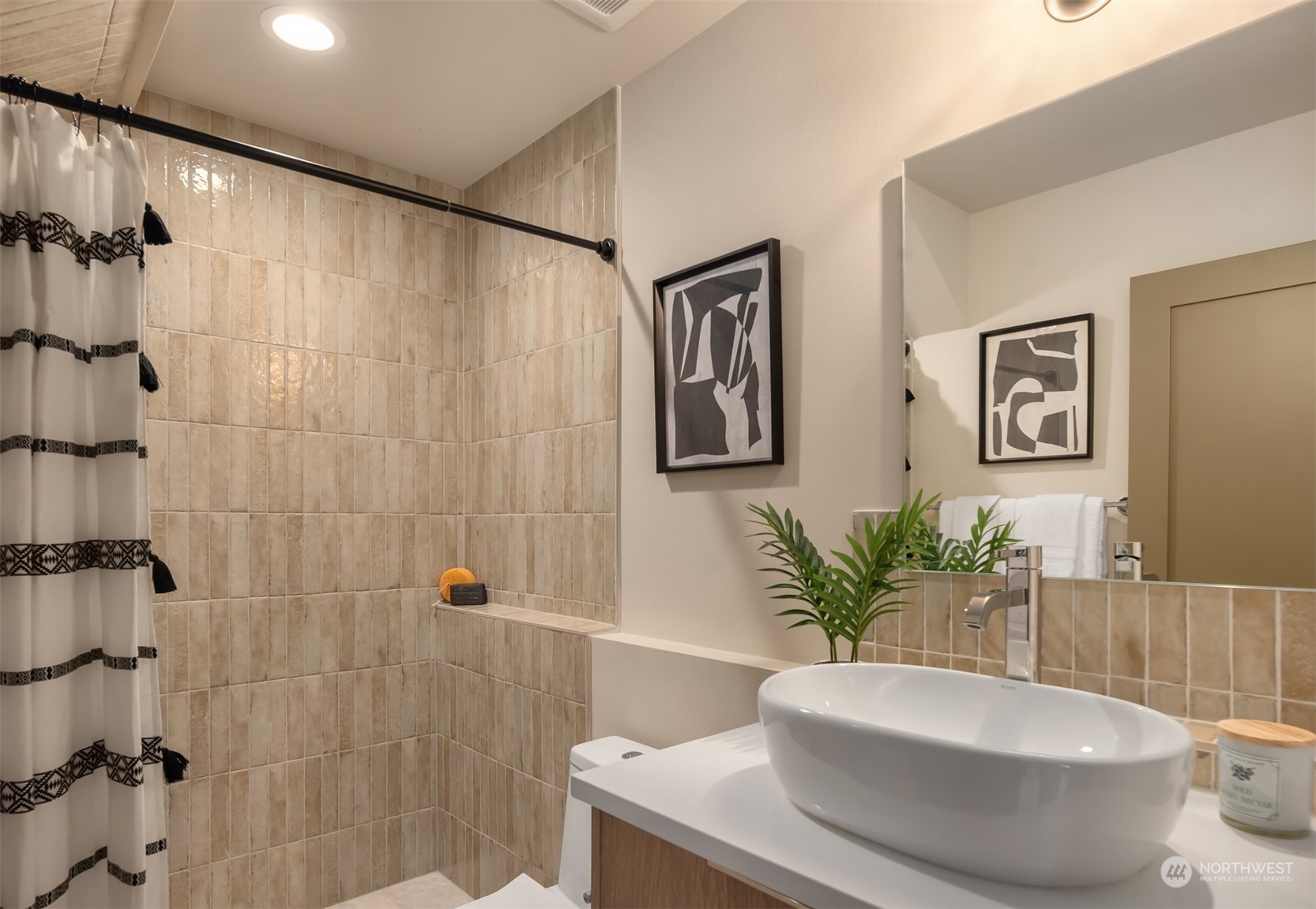 Bathroom with beige tiled shower, patterned curtain, white vessel sink, and wood-front vanity.