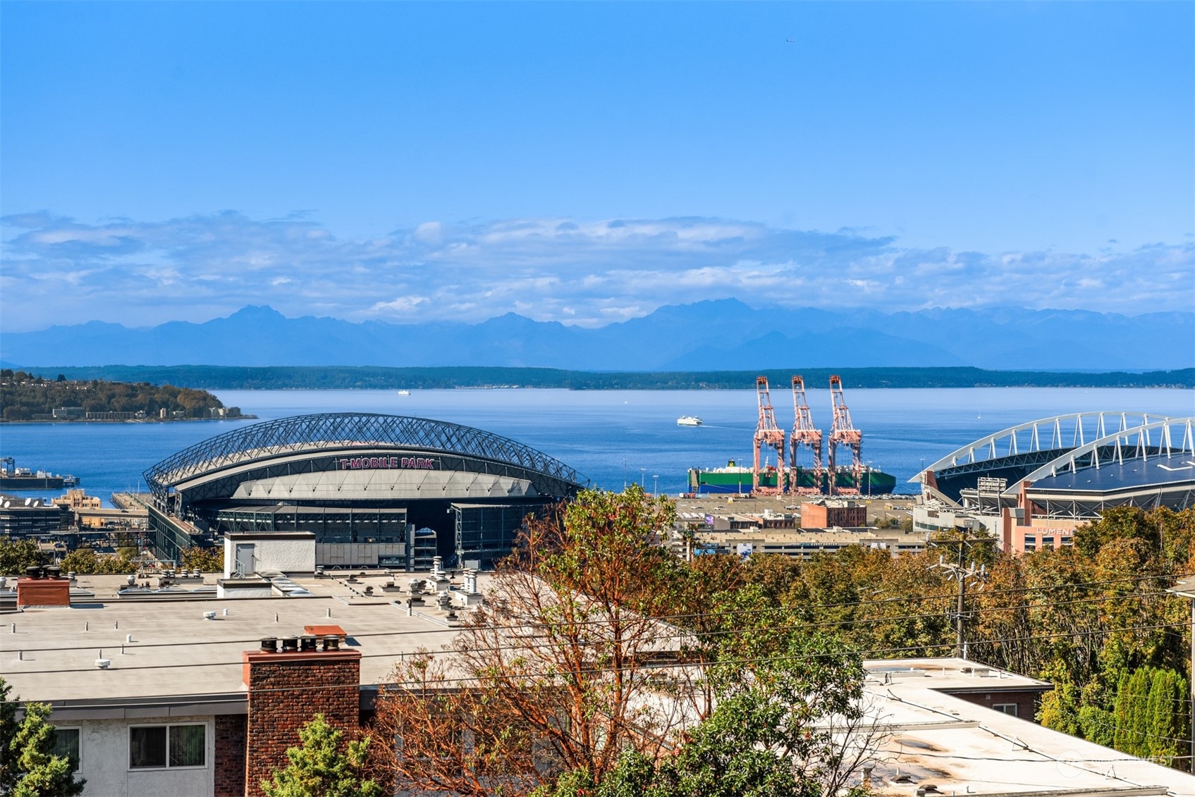 Seattle stadiums overlook Elliott Bay, port cranes, and distant mountains in an autumn panorama.