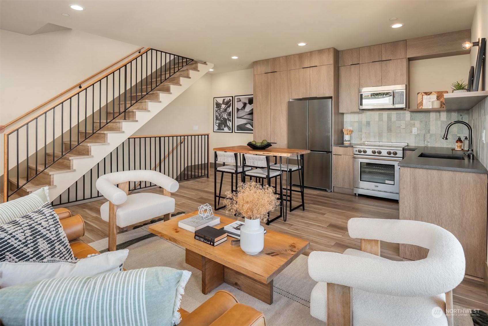 Open living room and kitchen with wood cabinetry, stainless steel appliances, and a staircase with black railings.