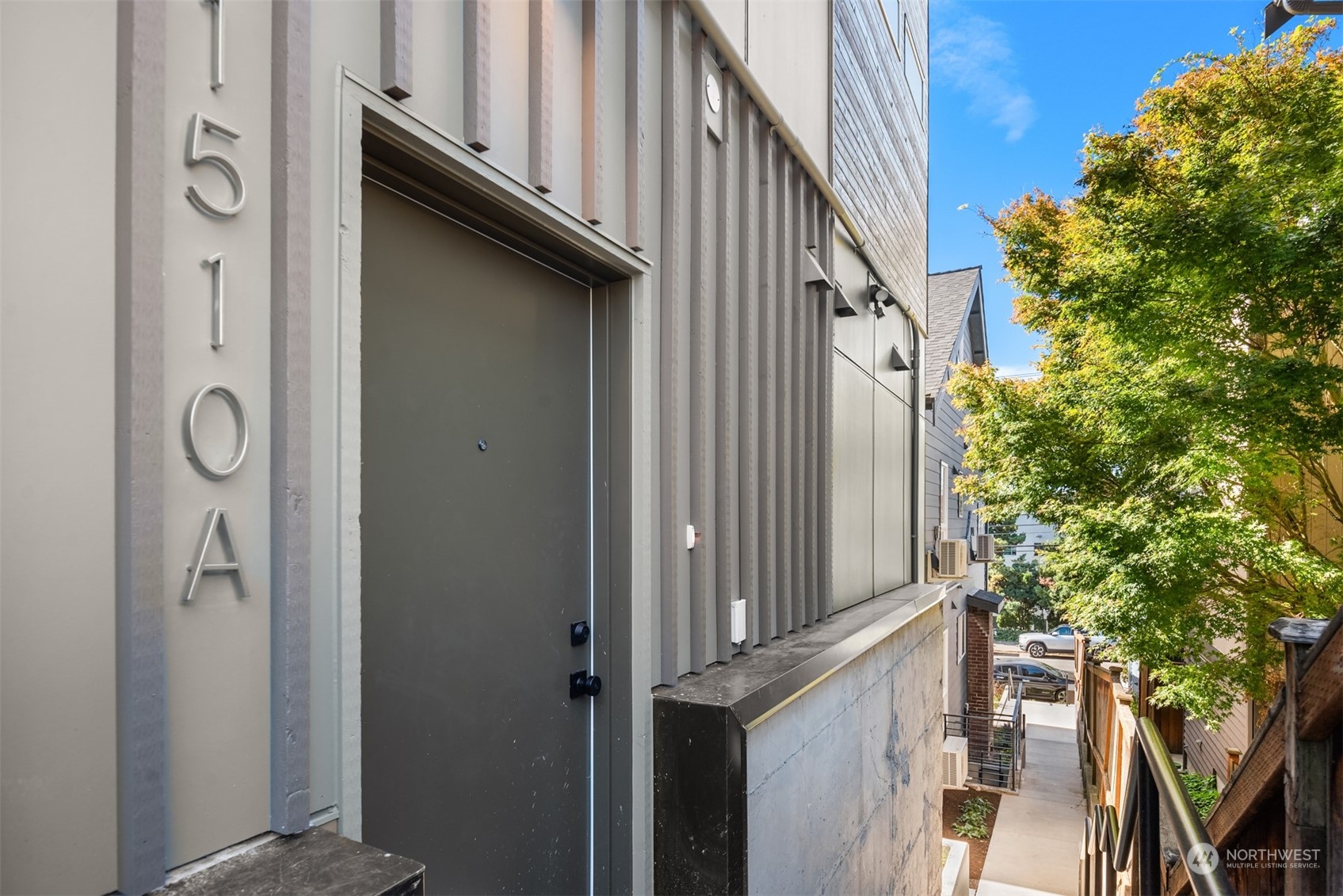 Modern gray home entrance with dark door, vertical facade details, and walkway beside autumn foliage.