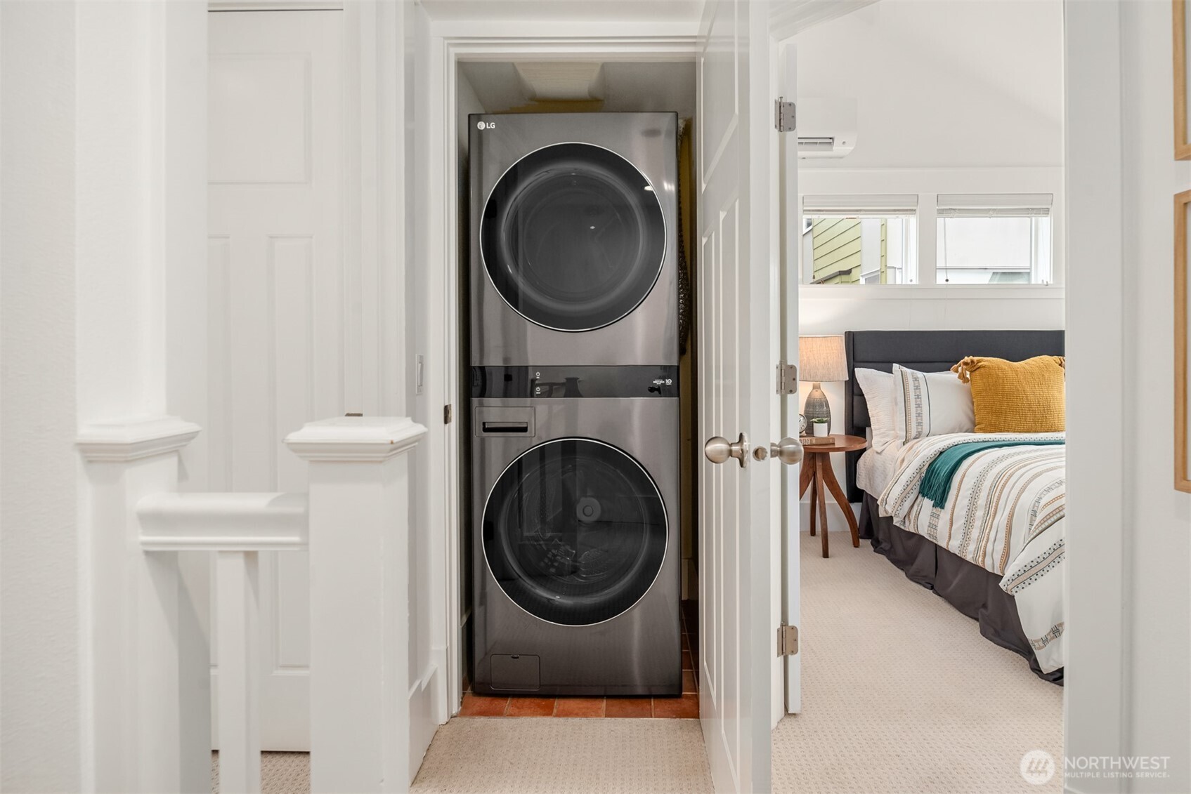 Stacked washer and dryer in a closet beside a bedroom with mustard-yellow accent pillows.