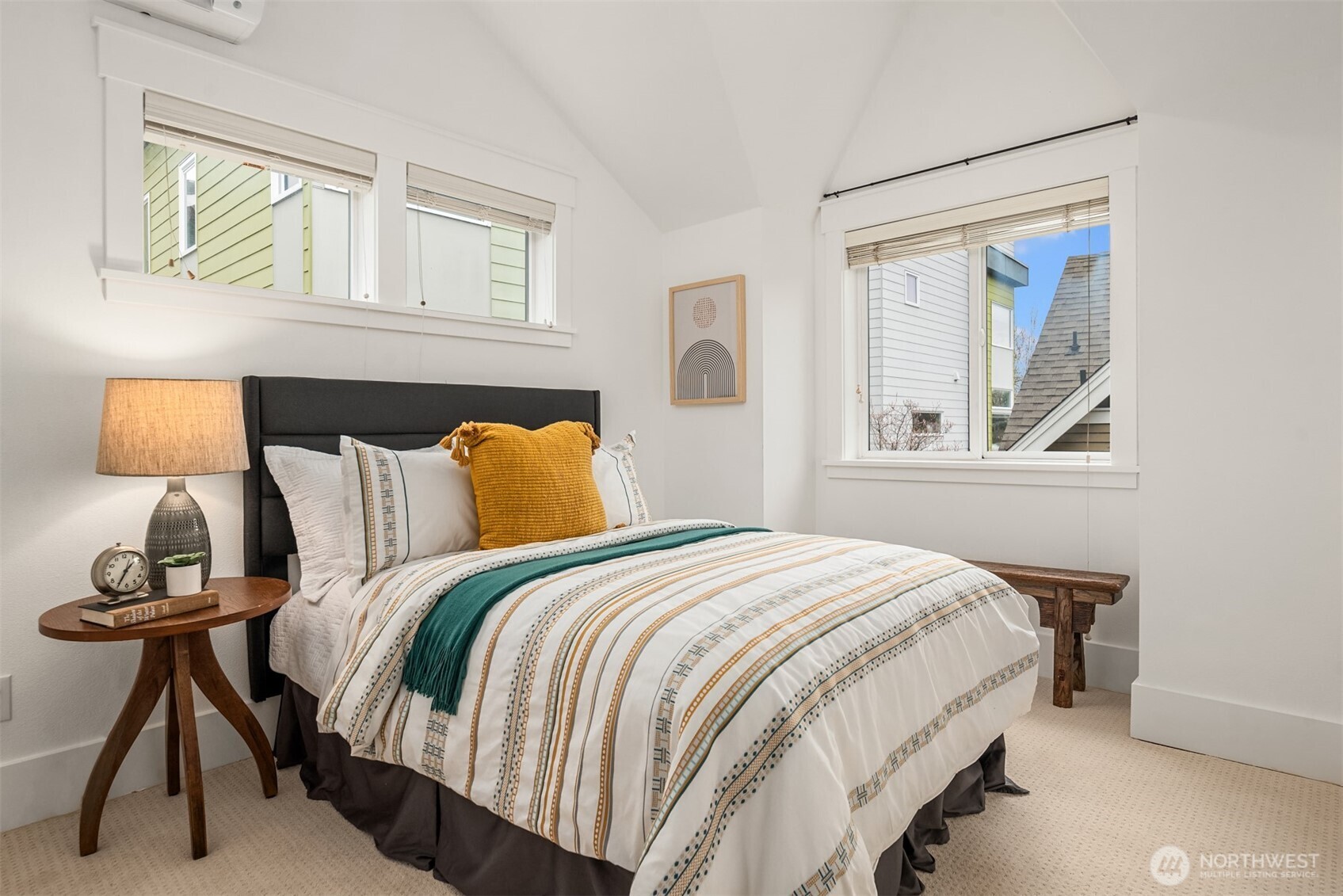 Staged bedroom with patterned bedding, teal and mustard pillows, and two daylight-filled windows.