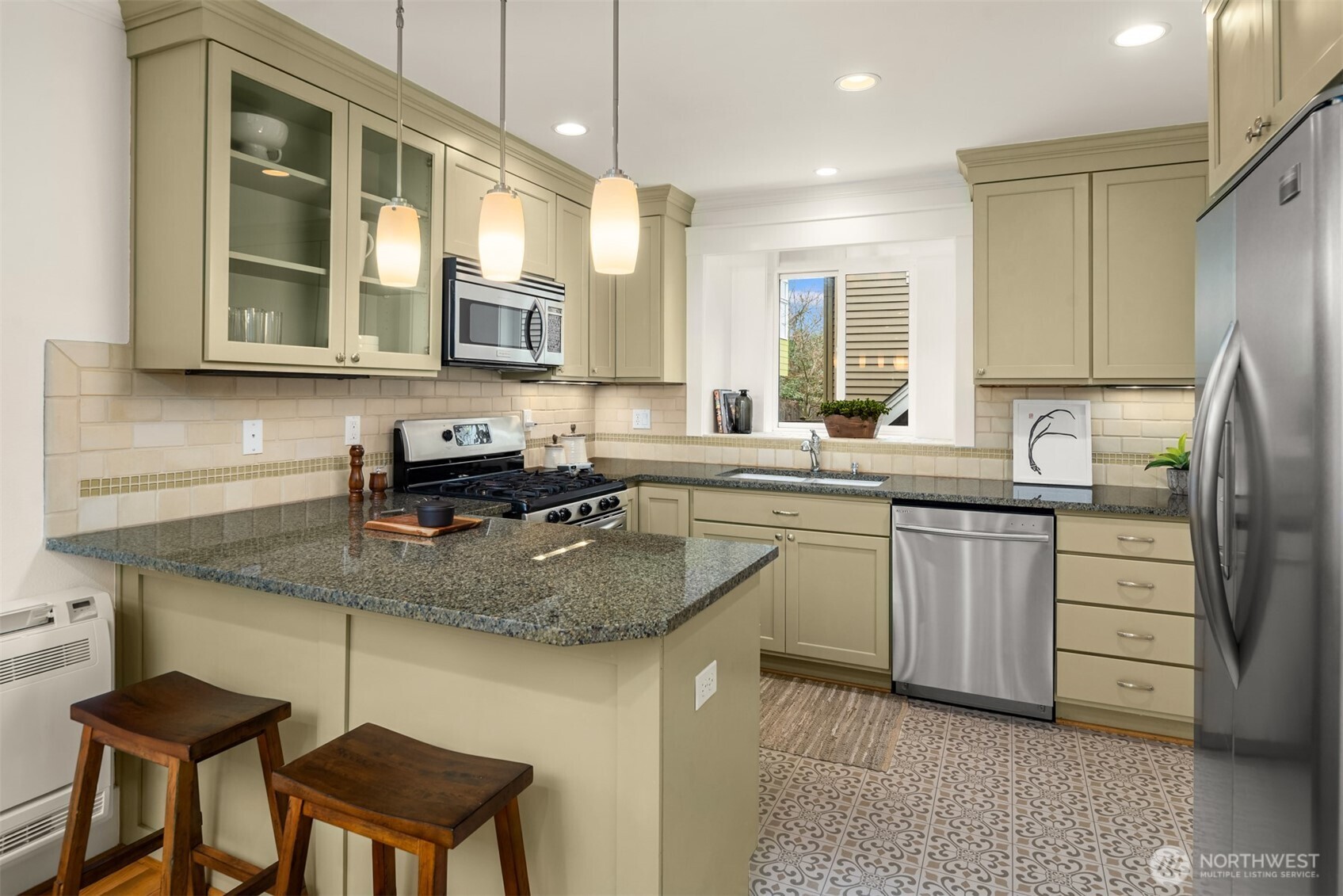 Kitchen with sage-green cabinets, granite counters, stainless steel appliances, and a breakfast bar with two stools.
