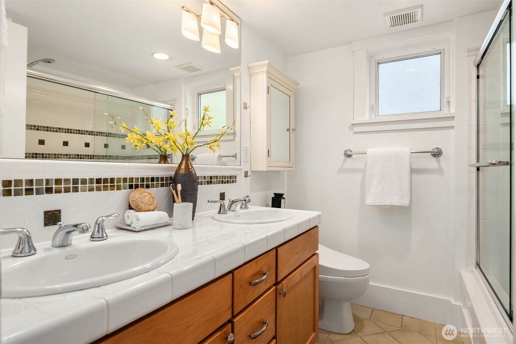 Bright bathroom with double sinks, a tiled vanity, warm wood cabinets, and a glass-enclosed shower.