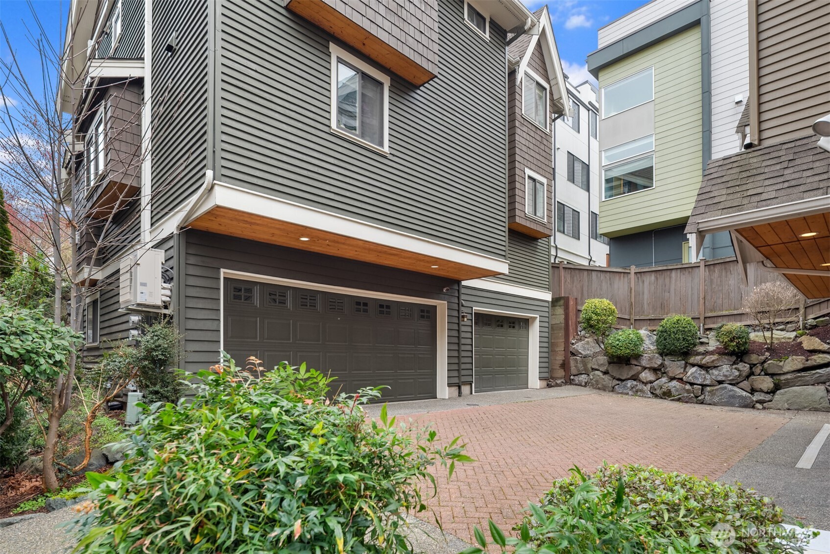 Townhouse exterior with two garage doors, a brick-paved driveway, and a stone retaining wall.