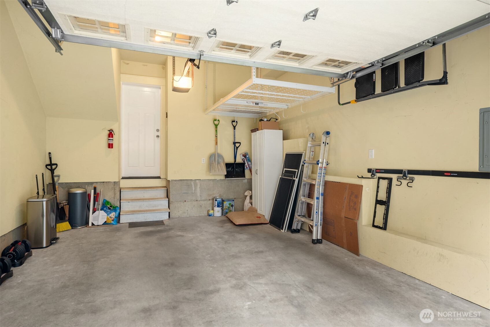 Garage with overhead storage racks, wall-mounted tool storage, ladders, and steps to a white door.