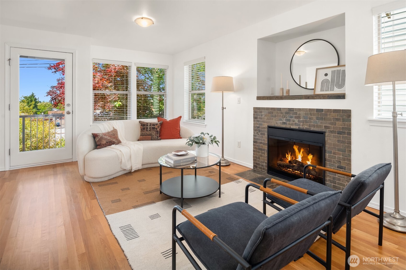 Staged living room with a lit brick fireplace, white sofa, dark lounge chairs, and glass door to a balcony.
