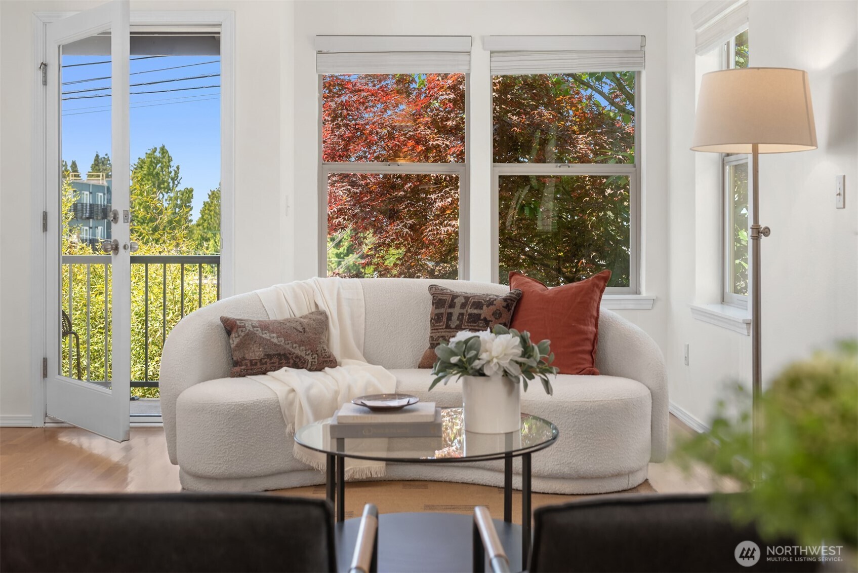 Staged living room with a curved white sofa, glass coffee table, and balcony doors overlooking autumn foliage.