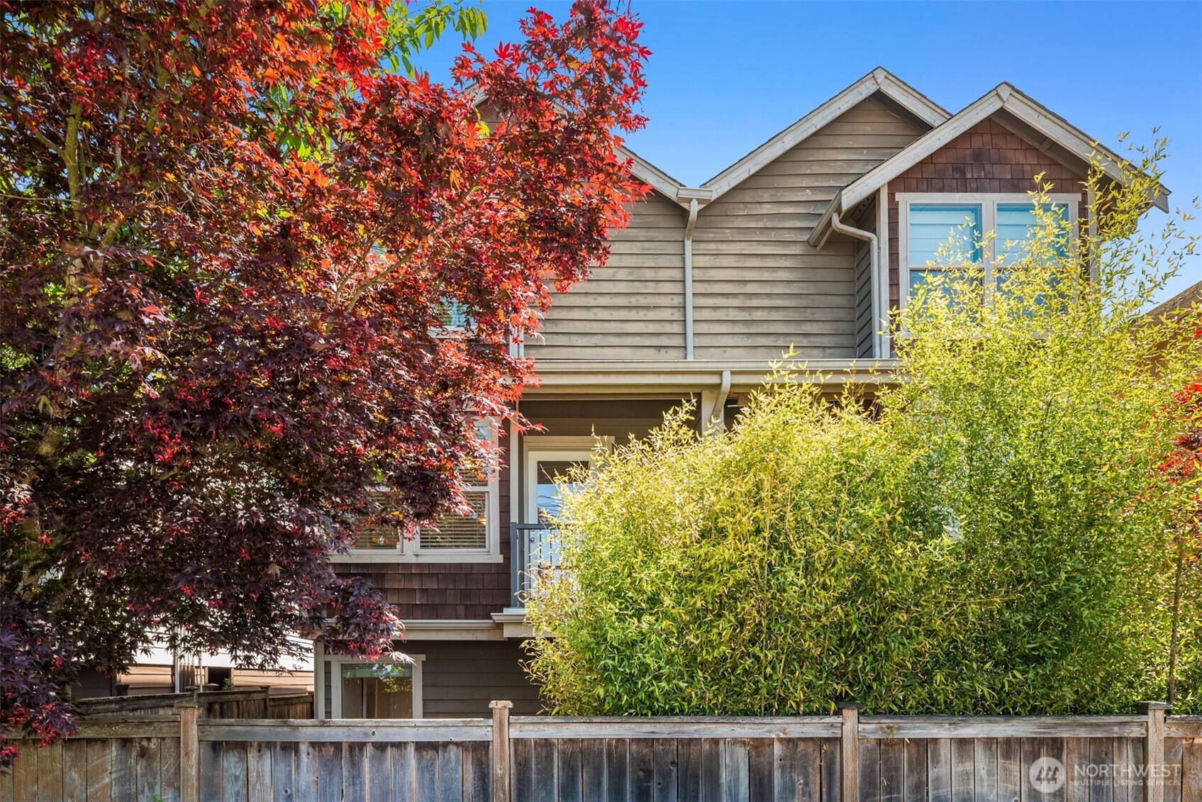 Multi-story gabled house behind a wooden fence, framed by red autumn foliage and tall shrubs.