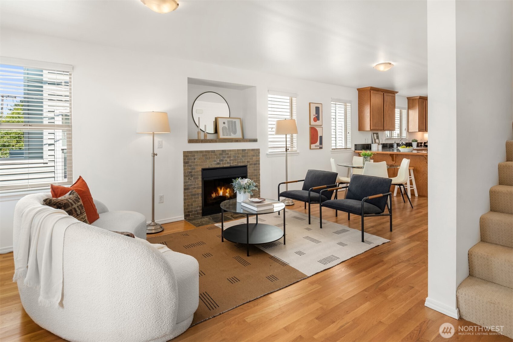 Staged living room with a tiled fireplace, white sofa, hardwood floors, and adjoining dining area and kitchen.