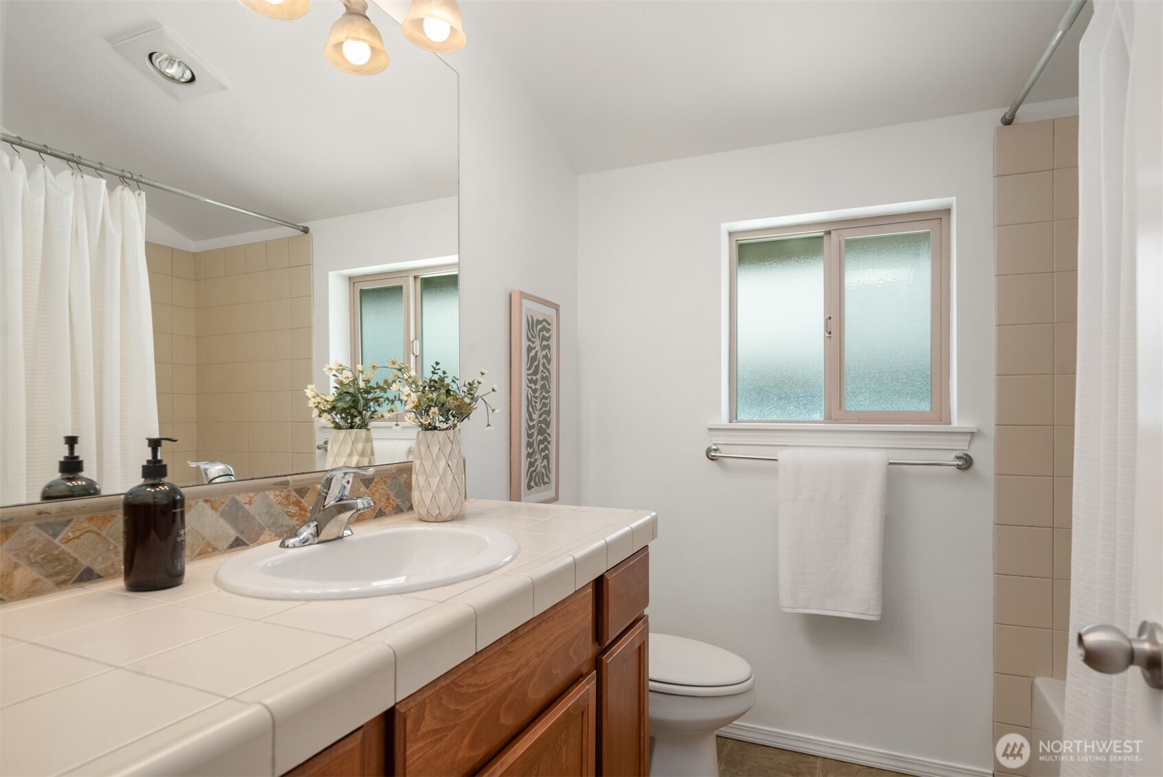 Bathroom with a tiled vanity, wooden cabinets, frosted window, and curtained tub and shower.